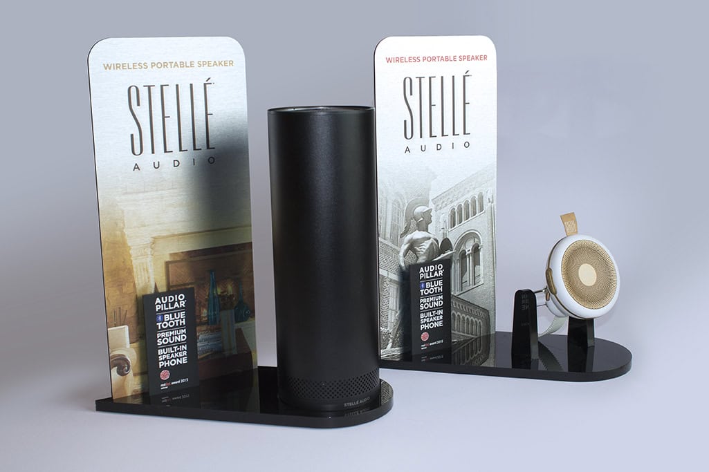 Display stand for Stellé Audio wireless portable speakers showcasing two different speaker models. One is a tall, cylindrical black speaker labeled "Audio Pillar" with features including Bluetooth, premium sound, and a built-in speakerphone. The other is a smaller, round white and gold speaker. The display boards behind the speakers feature the Stellé Audio branding and visuals of elegant interiors and architecture.