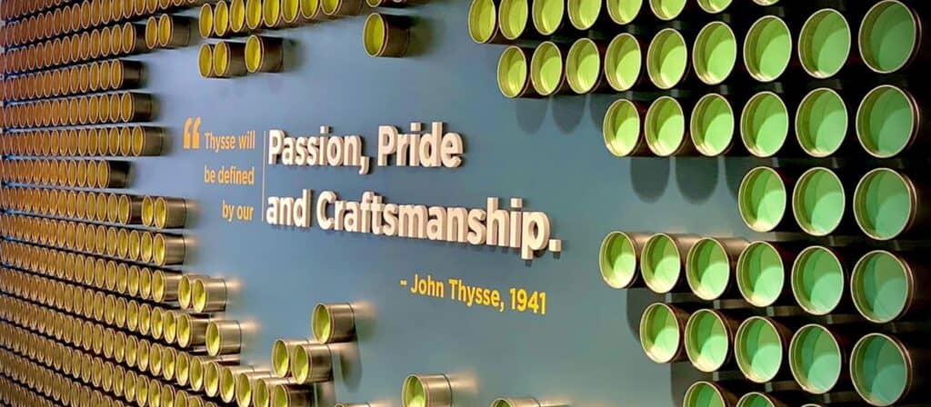 Decorative wall featuring rows of green ink cans with the quote 'Thysse will be defined by our Passion, Pride, and Craftsmanship' by John Thysse, 1941, at Thysse's Oregon, WI headquarters.