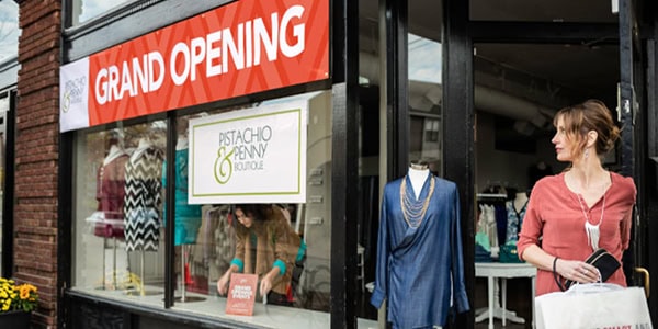 A boutique storefront with a bright red 'Grand Opening' banner above the window, and a customer walking out carrying a shopping bag.