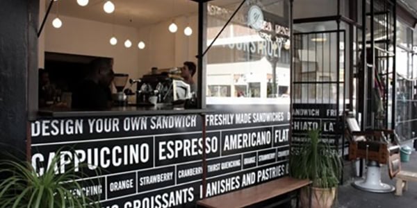 Black-and-white café window signage displaying menu items like espresso, sandwiches, and pastries, with a customer being served inside.