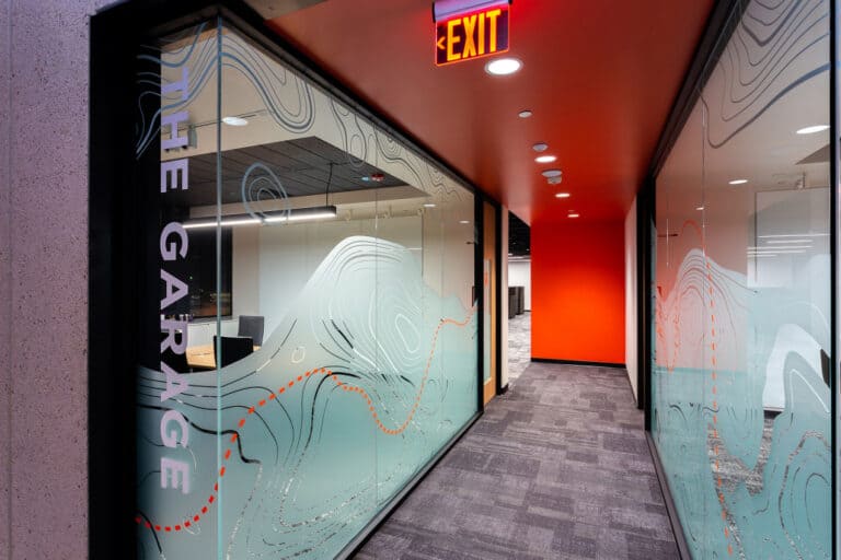 Bright hallway leading to 'The Garage' meeting space at Thysse's Oregon, WI headquarters, featuring frosted glass with topographic map designs and a bold red accent wall.