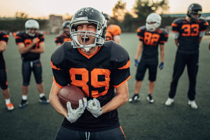 Football player wearing number 89 in full gear, celebrating with teammates in the background on the field.