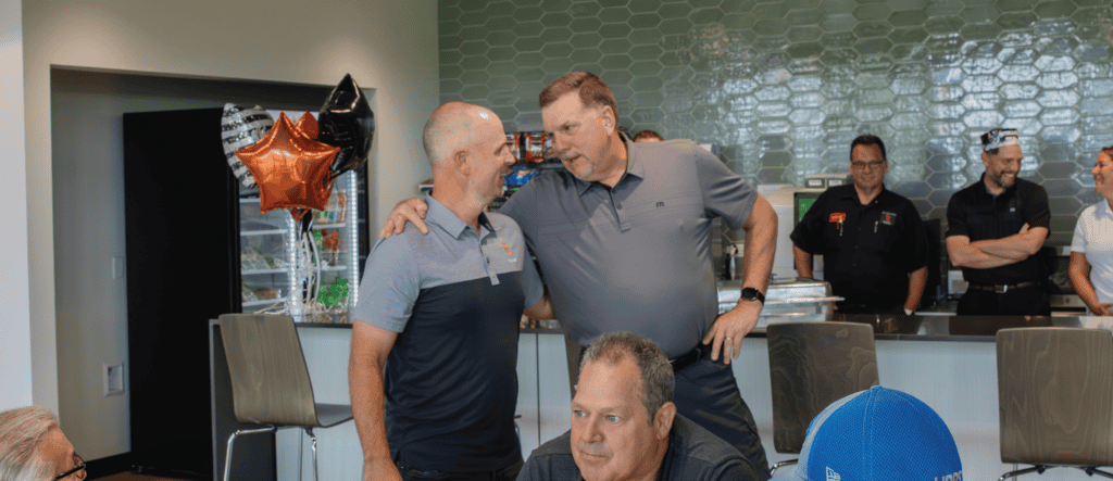 ason (left) and Dean (right) sharing a friendly moment during Jason's 30th work anniversary celebration. Jason is wearing a gray polo shirt, and Dean is wearing a gray collared shirt with his hand on Jason's shoulder. The background features balloons, snacks, and colleagues smiling and chatting near a counter.