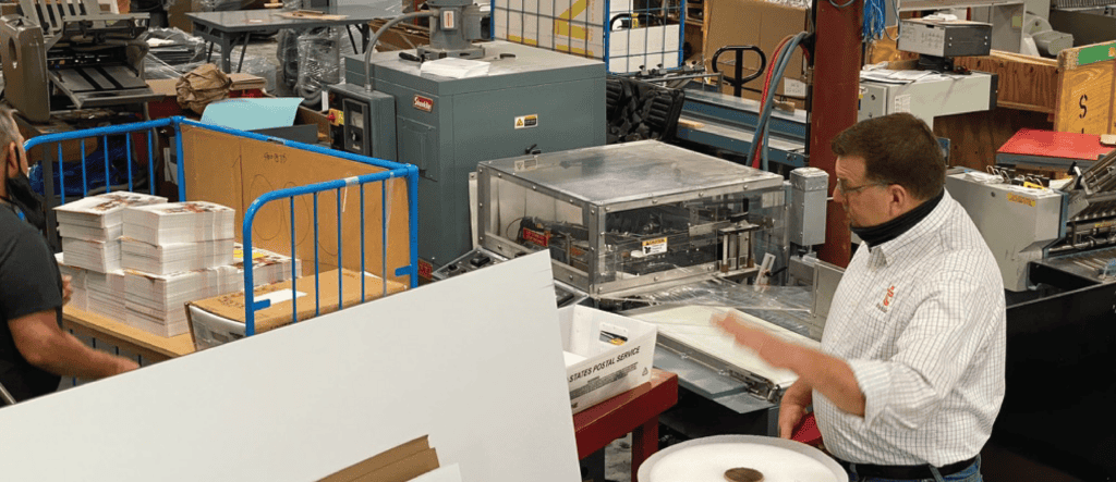 Dean speaking in Thysse's print shop, gesturing while explaining a process. He is wearing a light checkered shirt with the Thysse logo and a black neck gaiter. Surrounding him are printing machines, stacks of printed materials, and equipment, showcasing the busy shop environment.