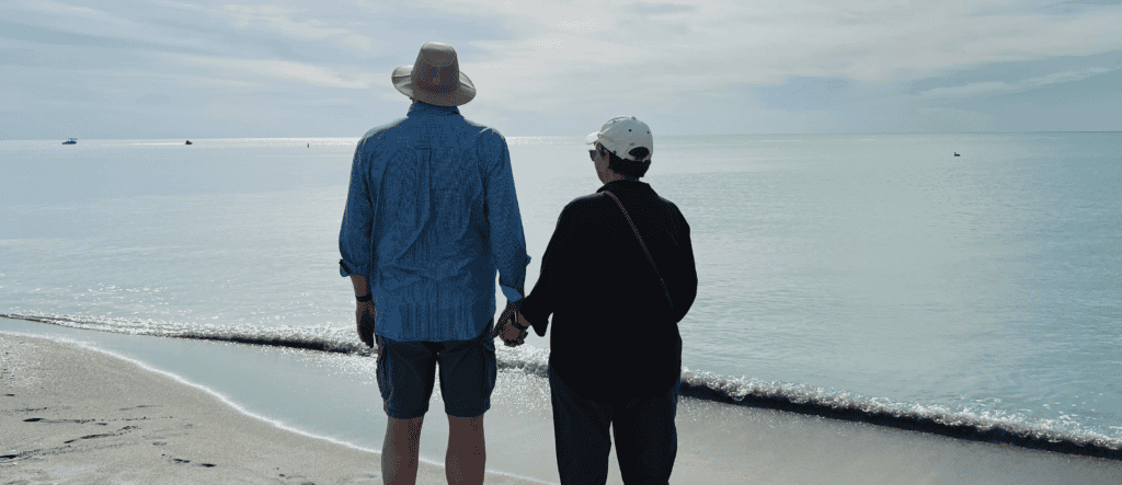 Dean and his wife, Dana, standing hand in hand on a serene beach, gazing out at the calm ocean under a lightly clouded sky. Dean is wearing a blue checkered shirt and a hat, while Dana is dressed in a black jacket and white cap, with gentle waves lapping at the shore.