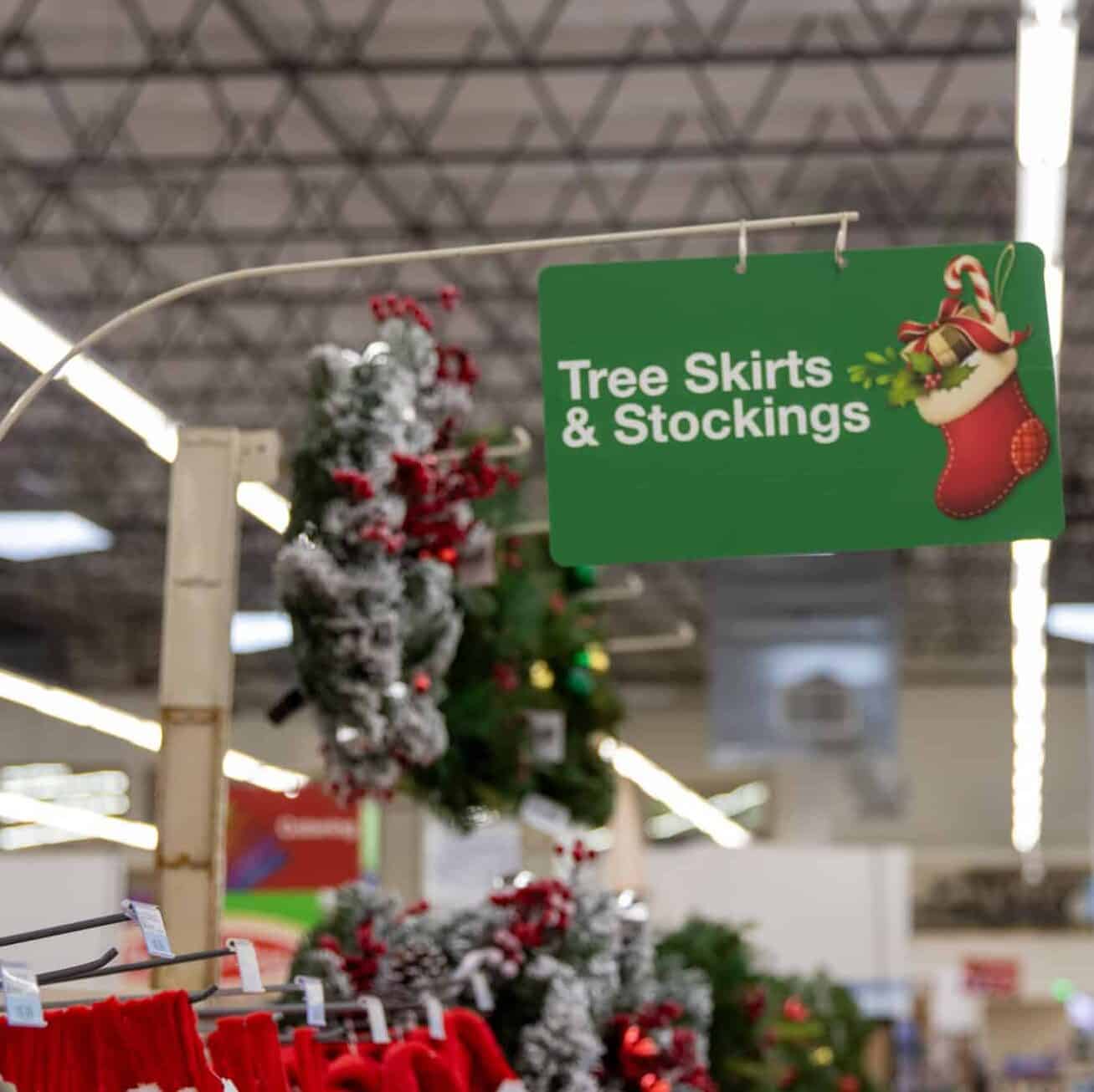 Sign hanging in an isle that reads Tree Skirts & Stockings, with an illustrated stocking filled with presents and candy canes. Wreaths and tops of stockings appear in the background, as well as the store ceiling.