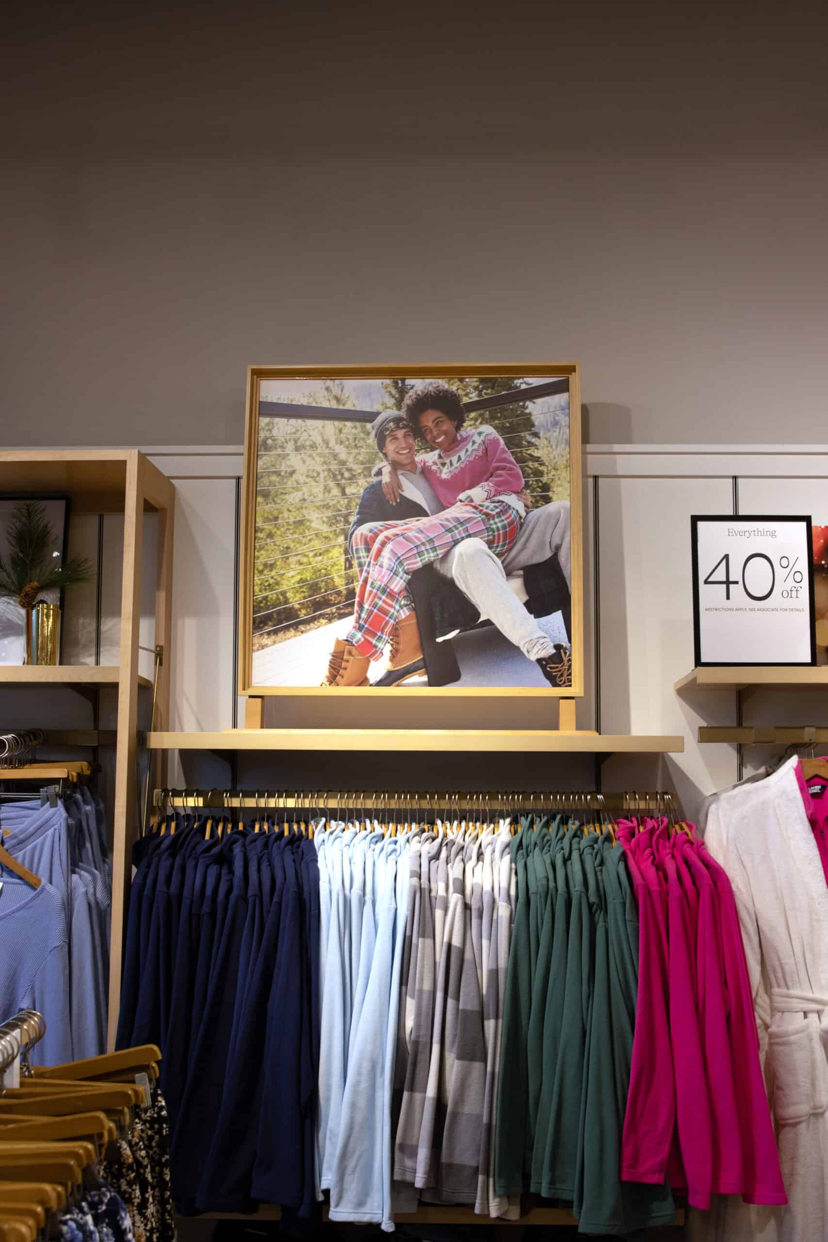 The image shows a Lands’ End store display featuring cozy and colorful women’s clothing. The centerpiece is a framed photo of two people smiling and embracing outdoors, wrapped in warm clothing and a plaid blanket. Beneath the photo, neatly hung fleece pullovers and robes in various colors, including navy, pastel blue, green, and pink, are organized on racks. A promotional sign to the right advertises "Everything 40% off," adding to the inviting shopping experience. The decor includes minimalist wooden shelving and a small holiday-themed arrangement on the left shelf.