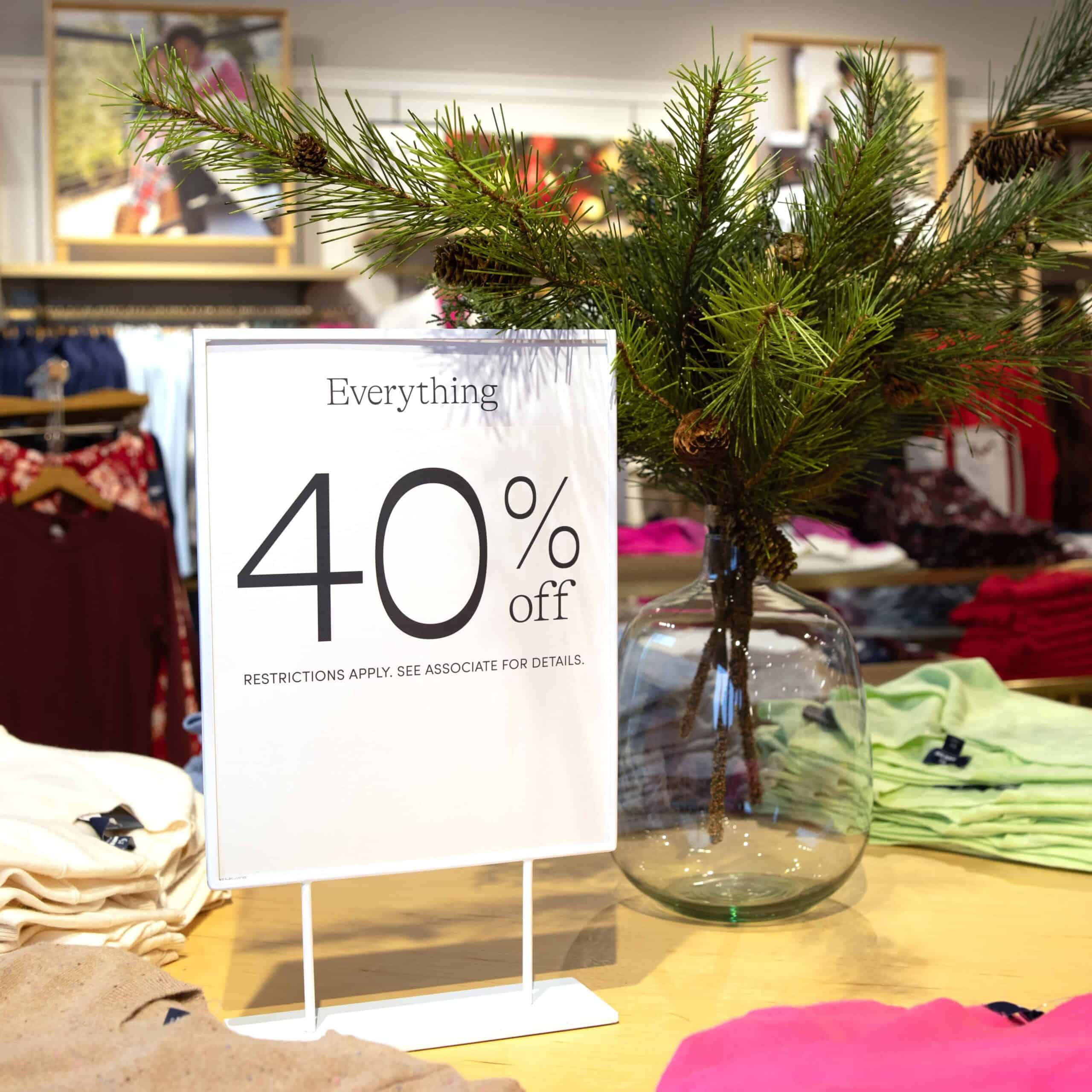 White sign with text that reads Everything 40% off. Restrictions apply. See associate for details. Conifer branches poking out of a clear, round glass vase. All on a maple table with clothes neatly stacked around. Clothes and lifestyle signage hanging behind in this retail environment.