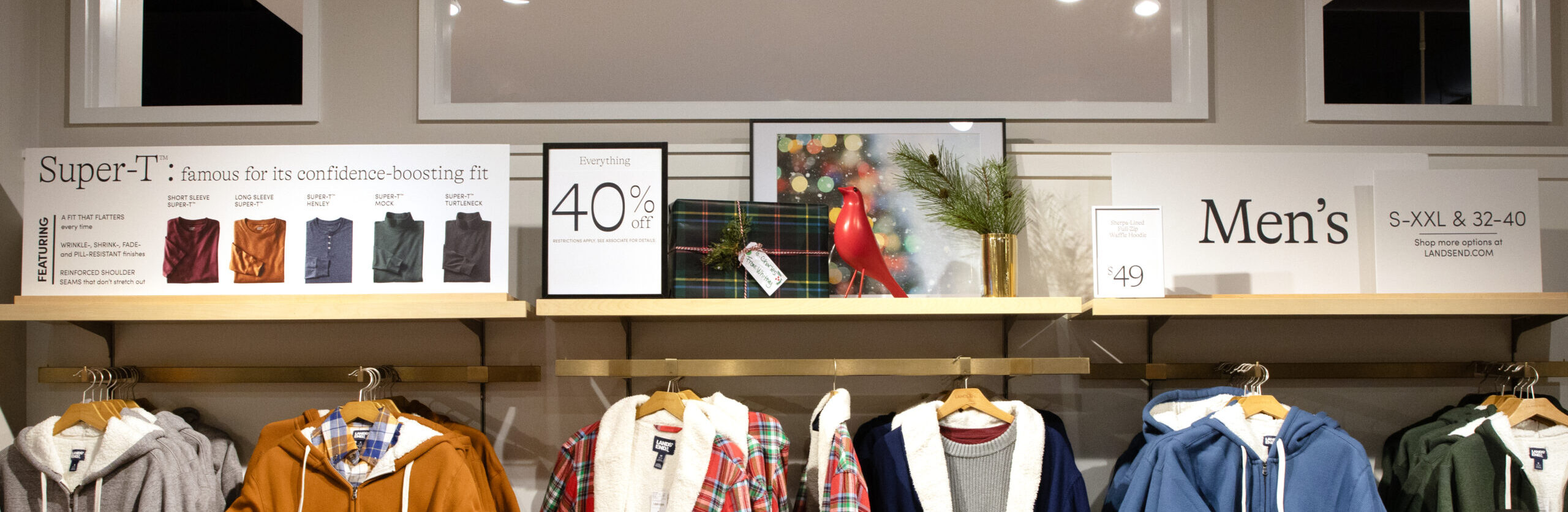 The image shows a men’s clothing display at the Lands’ End store in Middleton. The display features a variety of items, including hooded jackets, plaid robes, and sweaters. The top shelf has signs highlighting features like "Super-T: famous for its confidence-boosting fit," along with promotional messaging such as "Everything 40% off." Decorative elements, including a holiday-themed red bird figurine and wrapped gifts, enhance the seasonal shopping vibe. Additional signage directs customers to shop for more options online.