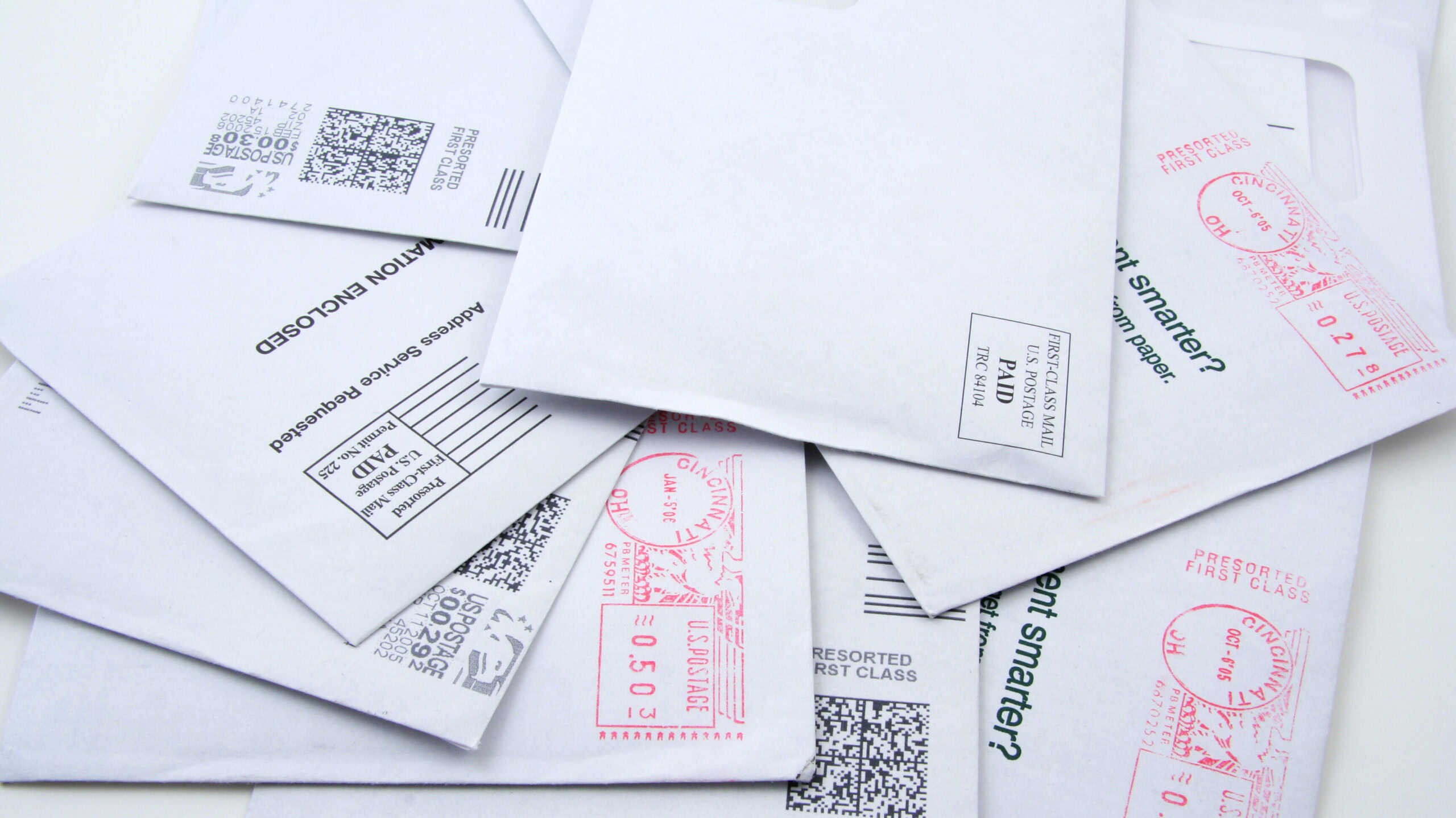 A pile of white USPS First-Class mail envelopes, each featuring various postage markings and barcodes.