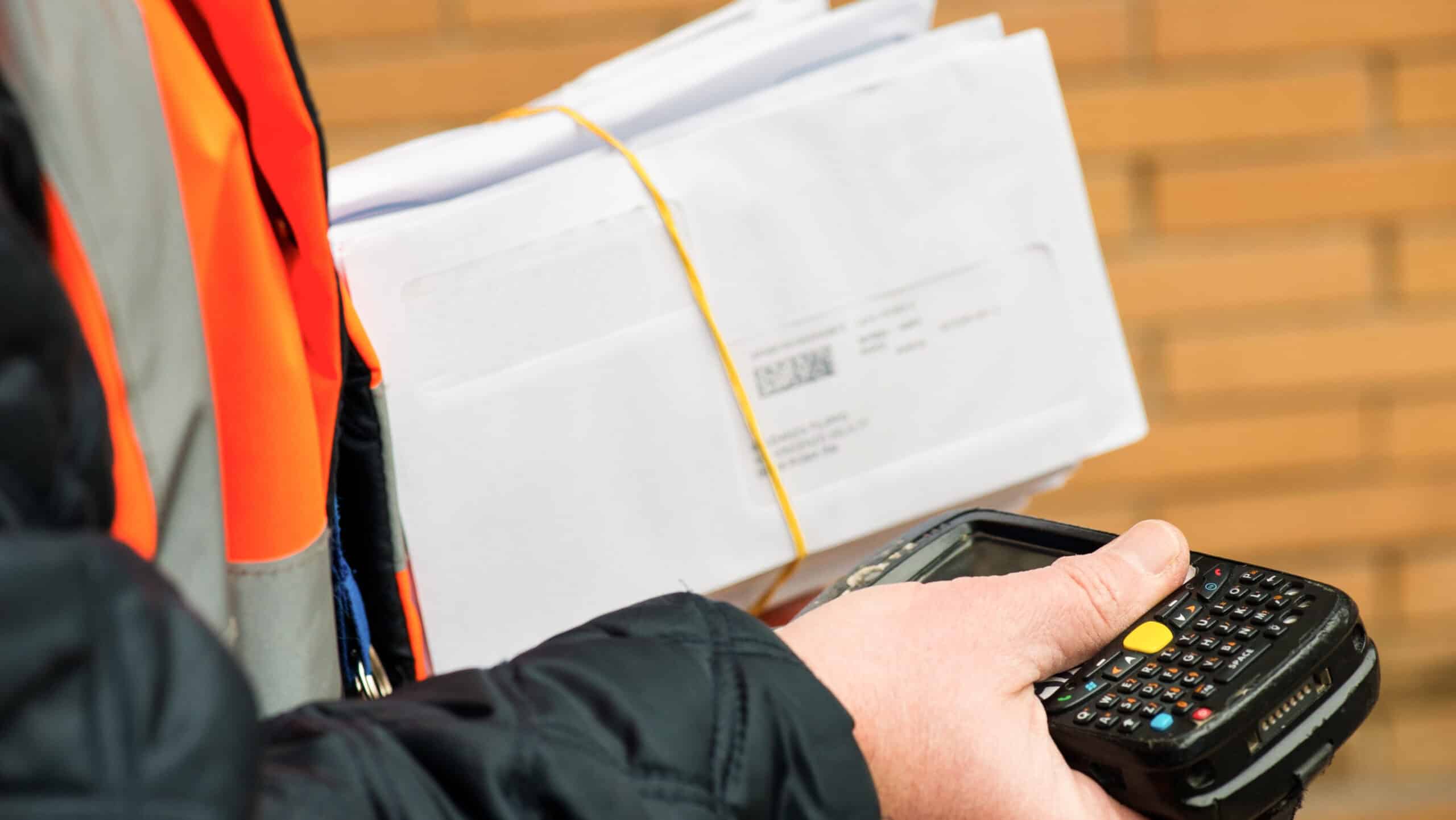 A postal worker holding a bundle of mail tied with a yellow rubber band, scanning with a handheld device against a brick wall background.