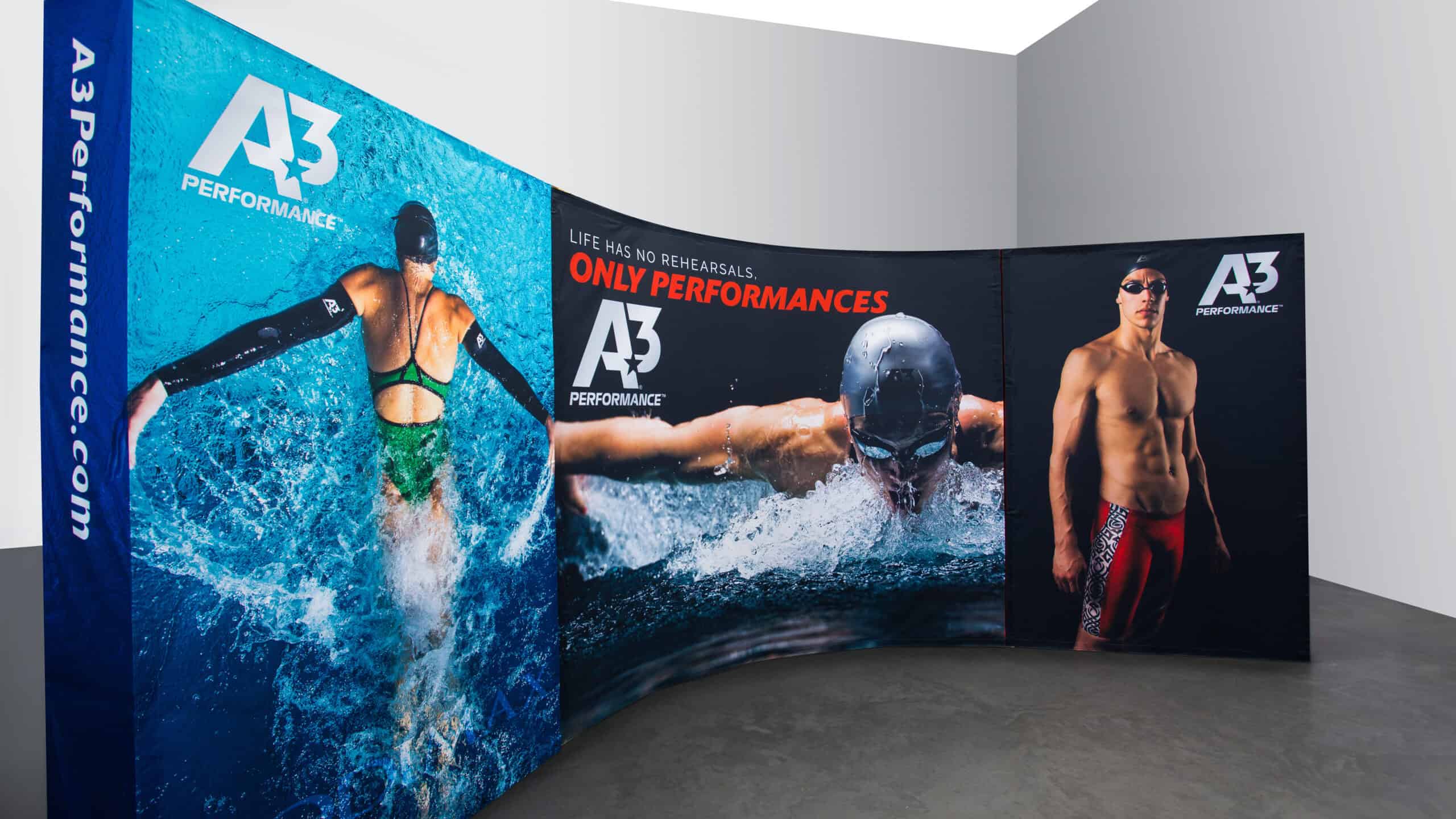 A dynamic, curved pop-up display for A3 Performance, featuring high-energy images of competitive swimmers in action. The bold graphics include the tagline "Life has no rehearsals. ONLY PERFORMANCES." in red and white lettering, reinforcing the brand's focus on elite swimwear and performance gear. The left panel showcases a backstroke swimmer cutting through the water, the center panel highlights a powerful butterfly stroke, and the right panel features a male swimmer in competition gear. The side panel prominently displays A3Performance.com in bold white lettering on a blue background.