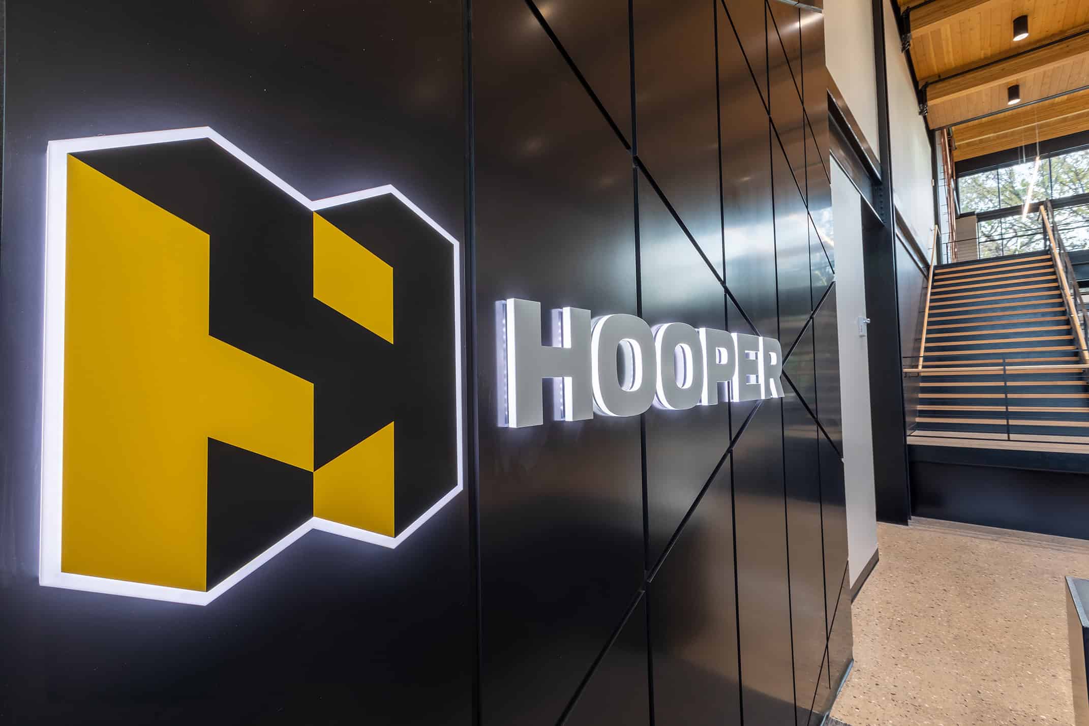 Hooper - Reception Signage A sleek, illuminated Hooper logo and lettering mounted on a dark, modern wall inside a corporate reception area. The bold yellow and black design contrasts with the contemporary architecture, featuring clean lines and a stairway leading to an upper level.