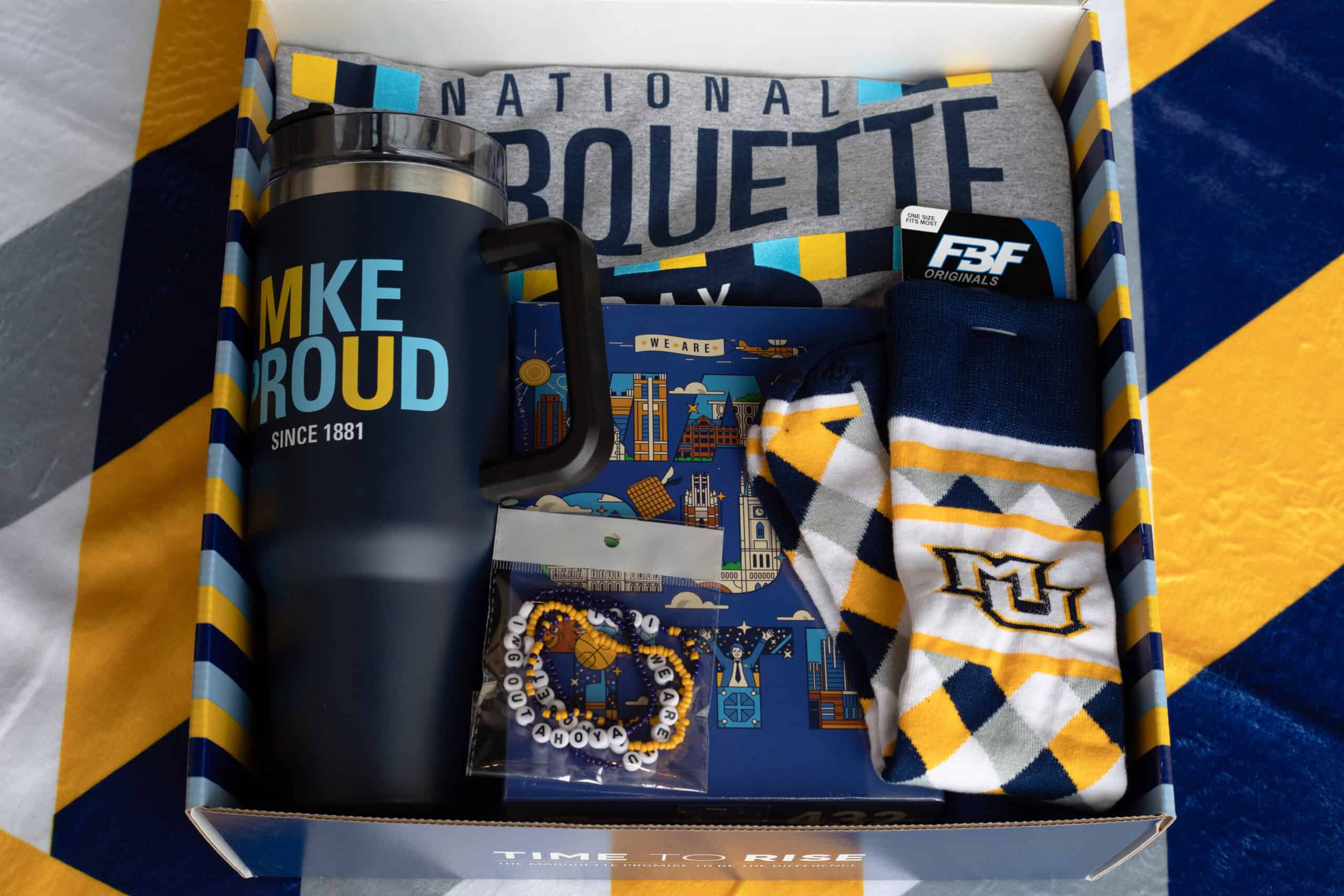 Marquette - Custom Gift Boxes A curated Marquette University gift box filled with branded merchandise, including a MKE Proud travel mug, socks, a T-shirt, a notebook, and beaded bracelets. The items are arranged in a custom-designed presentation box featuring the university’s blue and gold colors and geometric patterns.