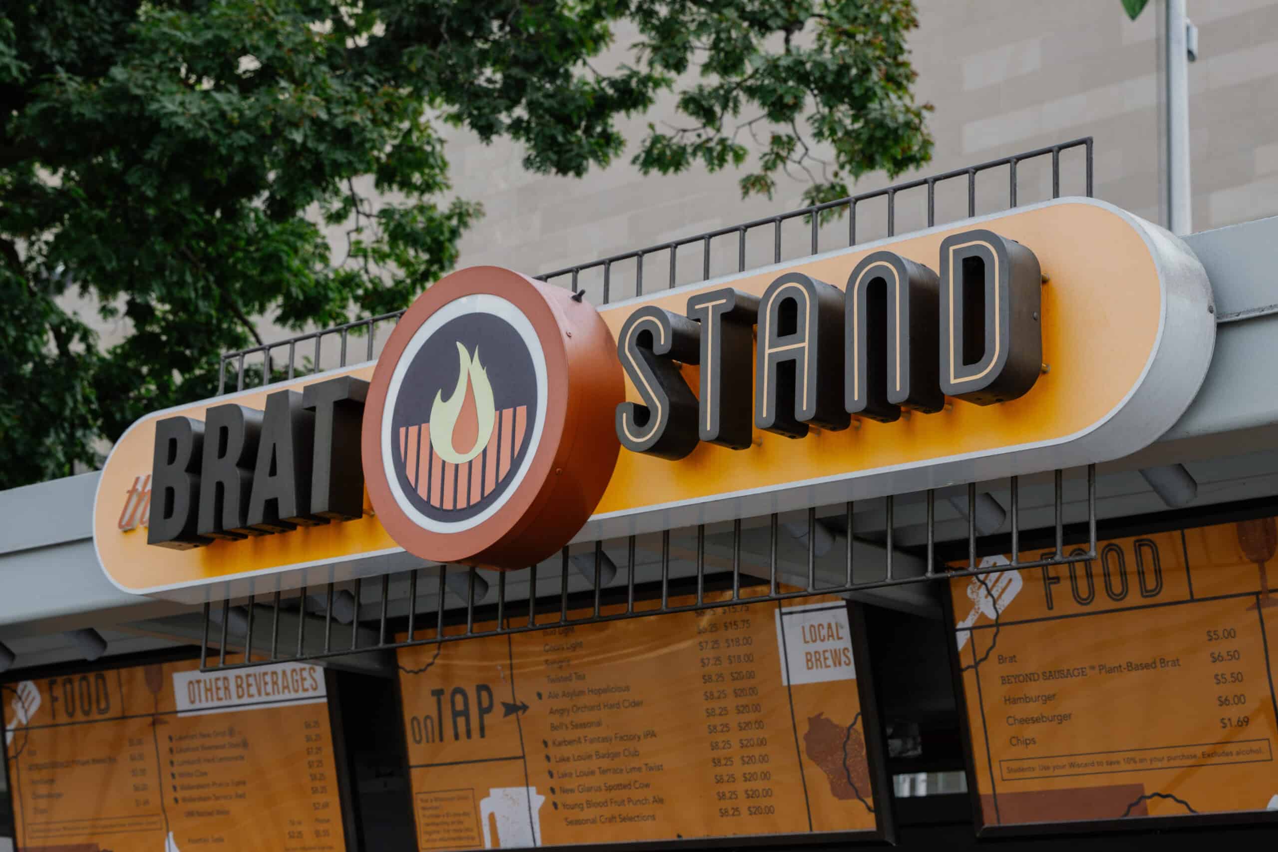 Memorial Union - Restaurant Signage A vibrant, three-dimensional restaurant sign for The Brat Stand, featuring bold typography and a circular grill flame icon. The yellow and black color scheme is paired with a detailed menu board below, set against an outdoor backdrop with trees and a neutral building facade.