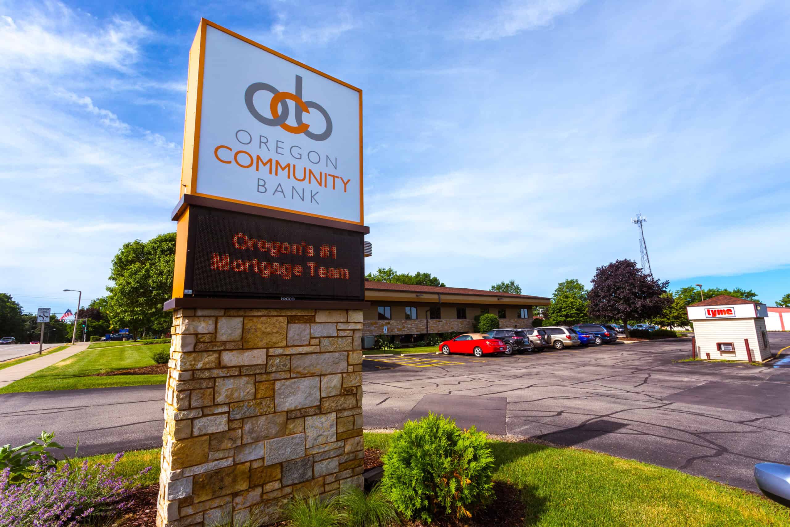 OCB - Exterior Messaging Sign A freestanding digital sign for Oregon Community Bank, featuring a combination of printed branding and an LED message board displaying mortgage team promotions. The sign is mounted on a stone base with a bright sky and commercial buildings in the background.