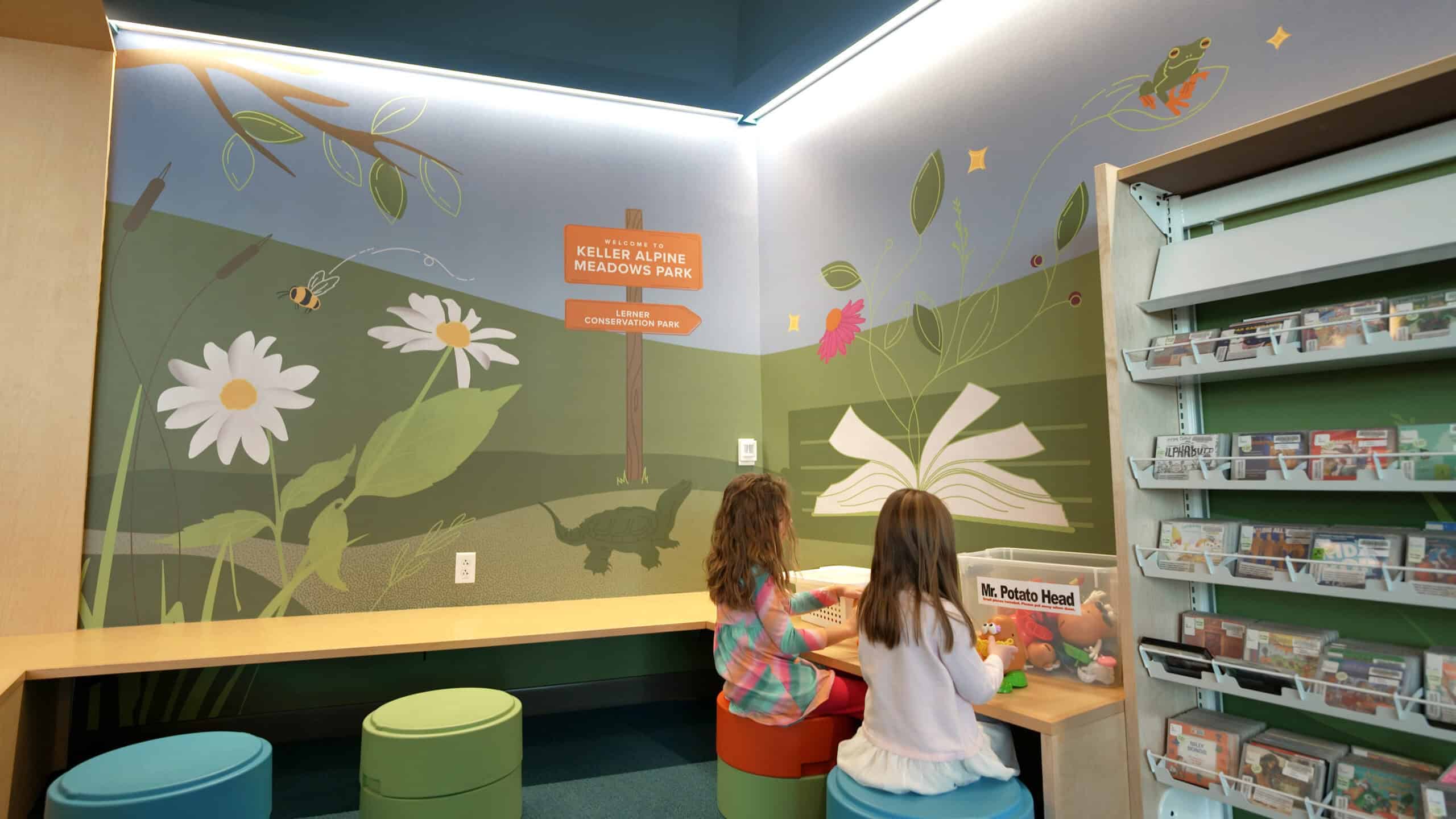 Oregon Public Library - Children's Nook A whimsical, nature-inspired reading nook at Oregon Public Library, featuring a hand-painted wall mural with rolling green hills, wildflowers, and illustrated animals. A signpost reading "Keller Alpine Meadows Park" adds a storytelling element. Two young children sit on colorful, round stools at a wooden activity bench, surrounded by books and educational materials.