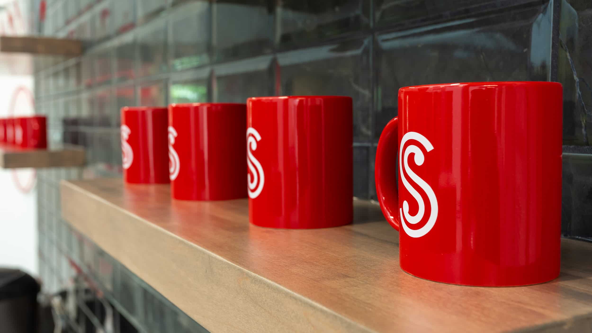 Society Insurance - Coffee Mugs A row of bold red ceramic coffee mugs featuring the Society Insurance logo in white. The mugs are neatly arranged on a wooden shelf against a backdrop of glossy dark green subway tiles, creating a clean, modern aesthetic.