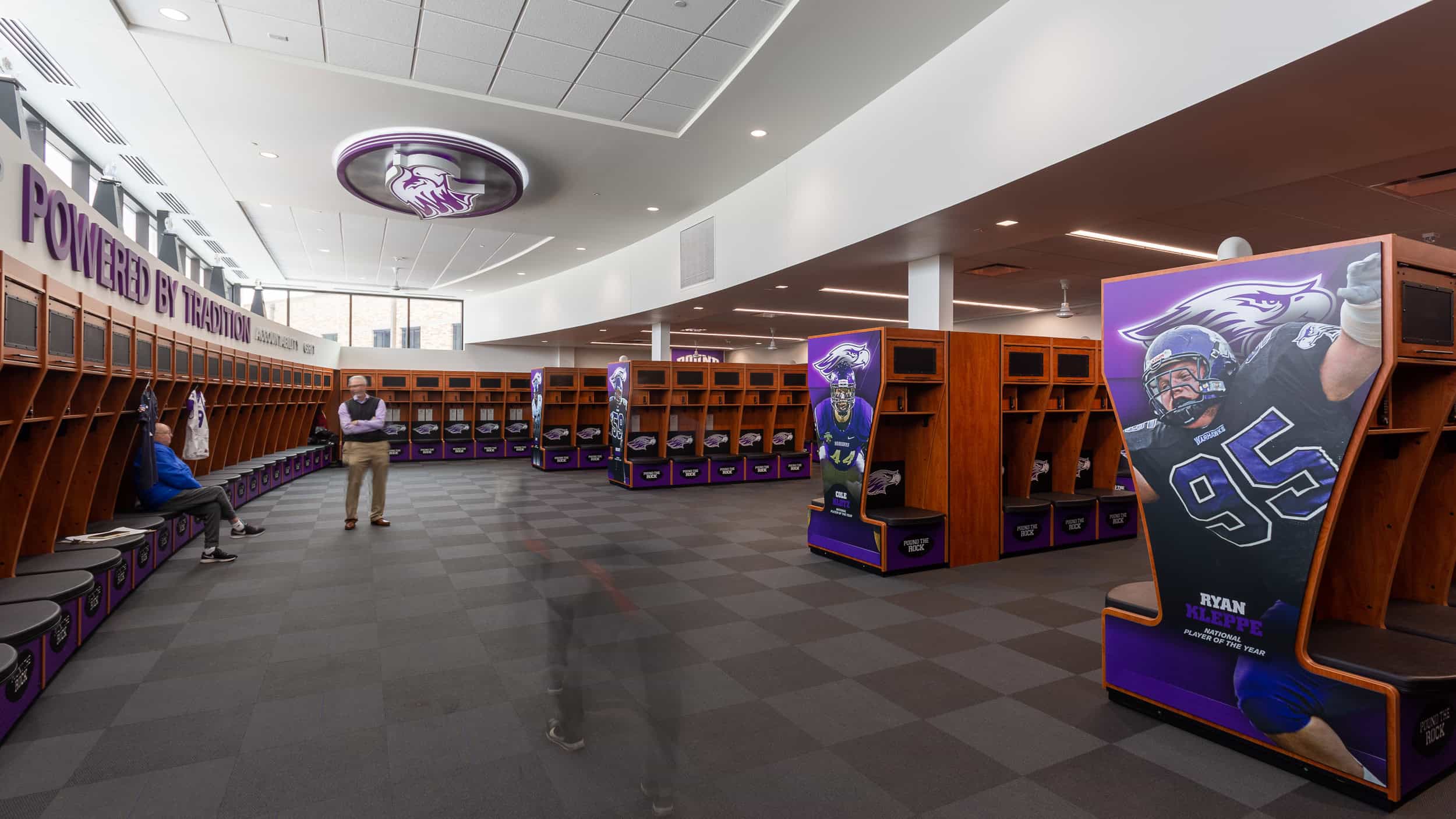 UW Whitewater - Football Locker Room Branding A fully branded locker room for UW-Whitewater Football, featuring customized lockers with large-scale player graphics and a bold “Powered by Tradition” message on the curved ceiling. The space includes wooden lockers with purple and black team branding, showcasing the team’s pride and legacy. A visitor in motion creates a subtle blur, adding to the sense of movement in the space.