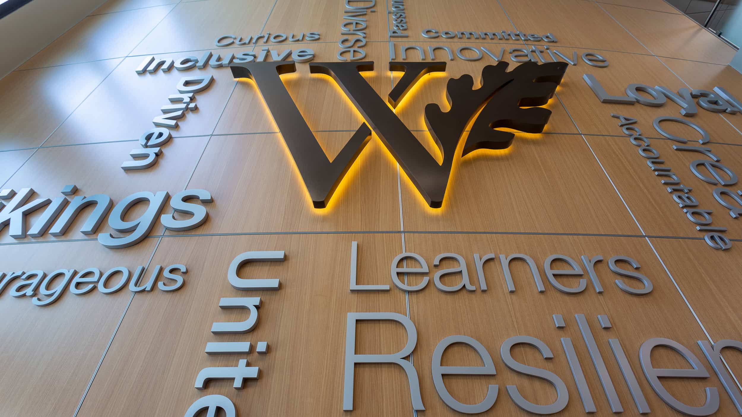 West Valley - Dimensional Lettering A close-up view of dimensional wall lettering in a modern educational or corporate space. The large metallic "W" is illuminated with soft backlighting, surrounded by words like "Resilient," "Learners," and "Innovative," emphasizing values and identity in a sophisticated, warm wood-paneled setting.