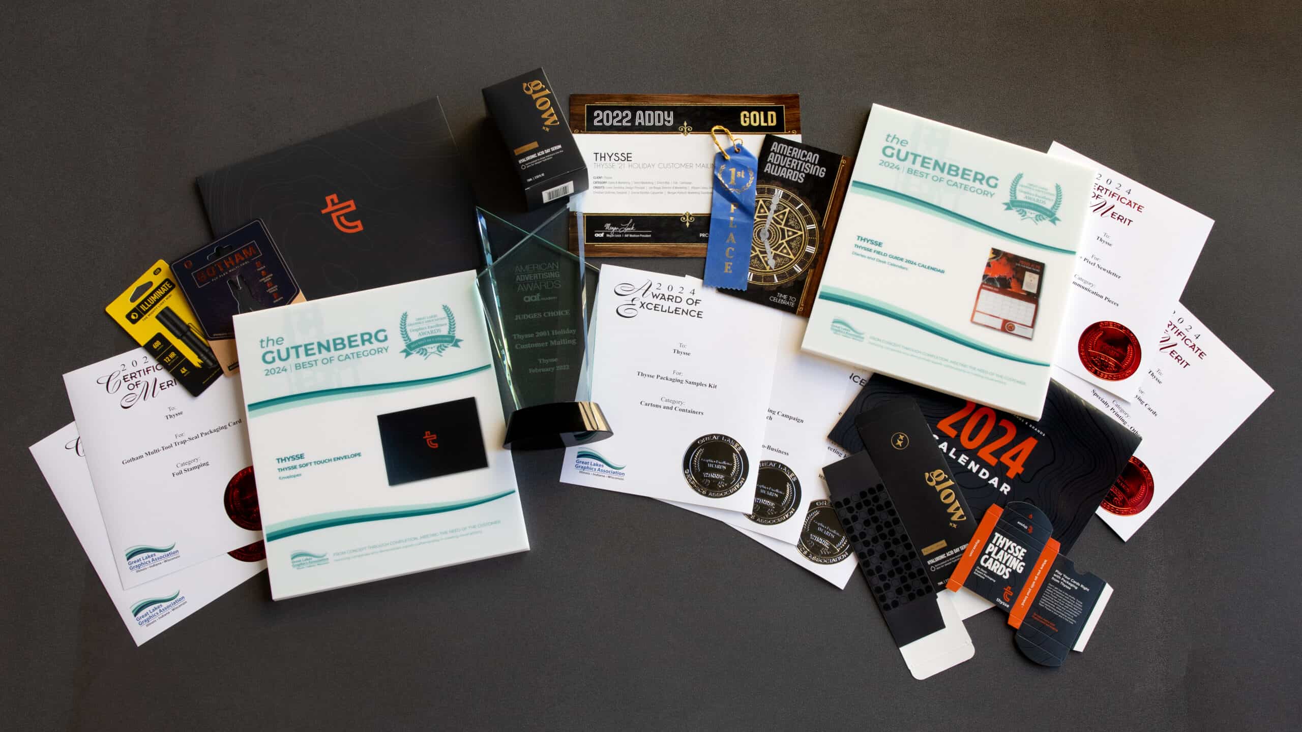 An assortment of Thysse's award-winning design projects and certificates displayed on a dark surface. The collection includes multiple Gutenberg 2024 'Best of Category' awards, an American Advertising Awards (ADDY) Gold trophy, an Award of Excellence plaque, and various certificates of merit. Also featured are custom-designed packaging samples, a 2024 calendar, branded playing cards, and other printed materials, showcasing Thysse's expertise in print and packaging design.