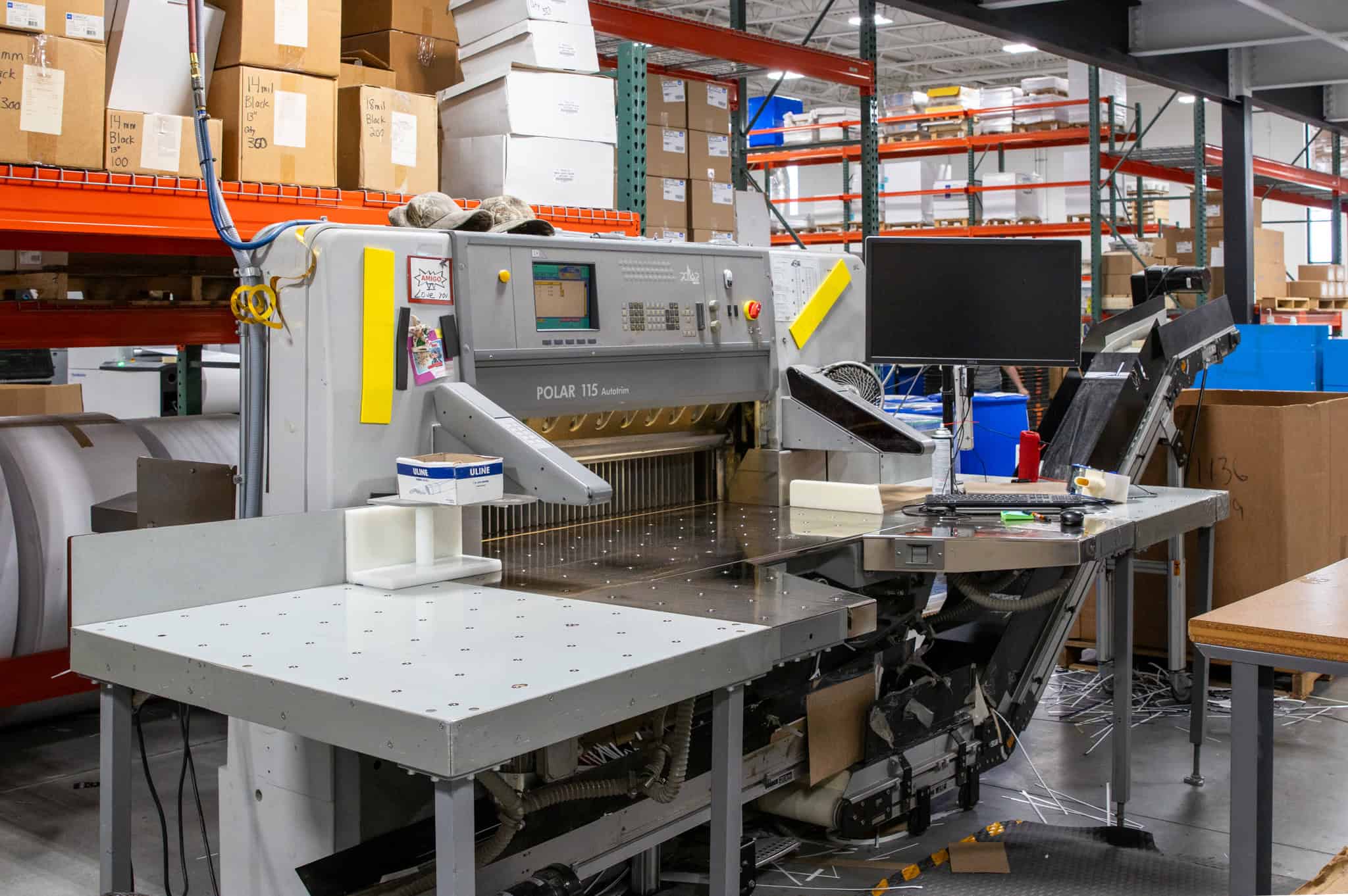 POLAR 115 Autotrim paper cutting machine in a print production facility, surrounded by shelving with boxed materials and a workstation with a monitor.
