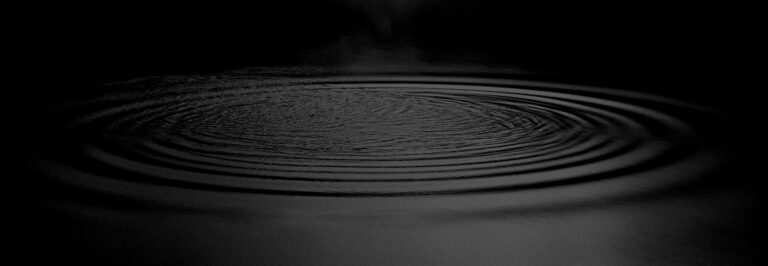 Concentric ripples spread across a dark, still body of water, creating a subtle pattern under low light.