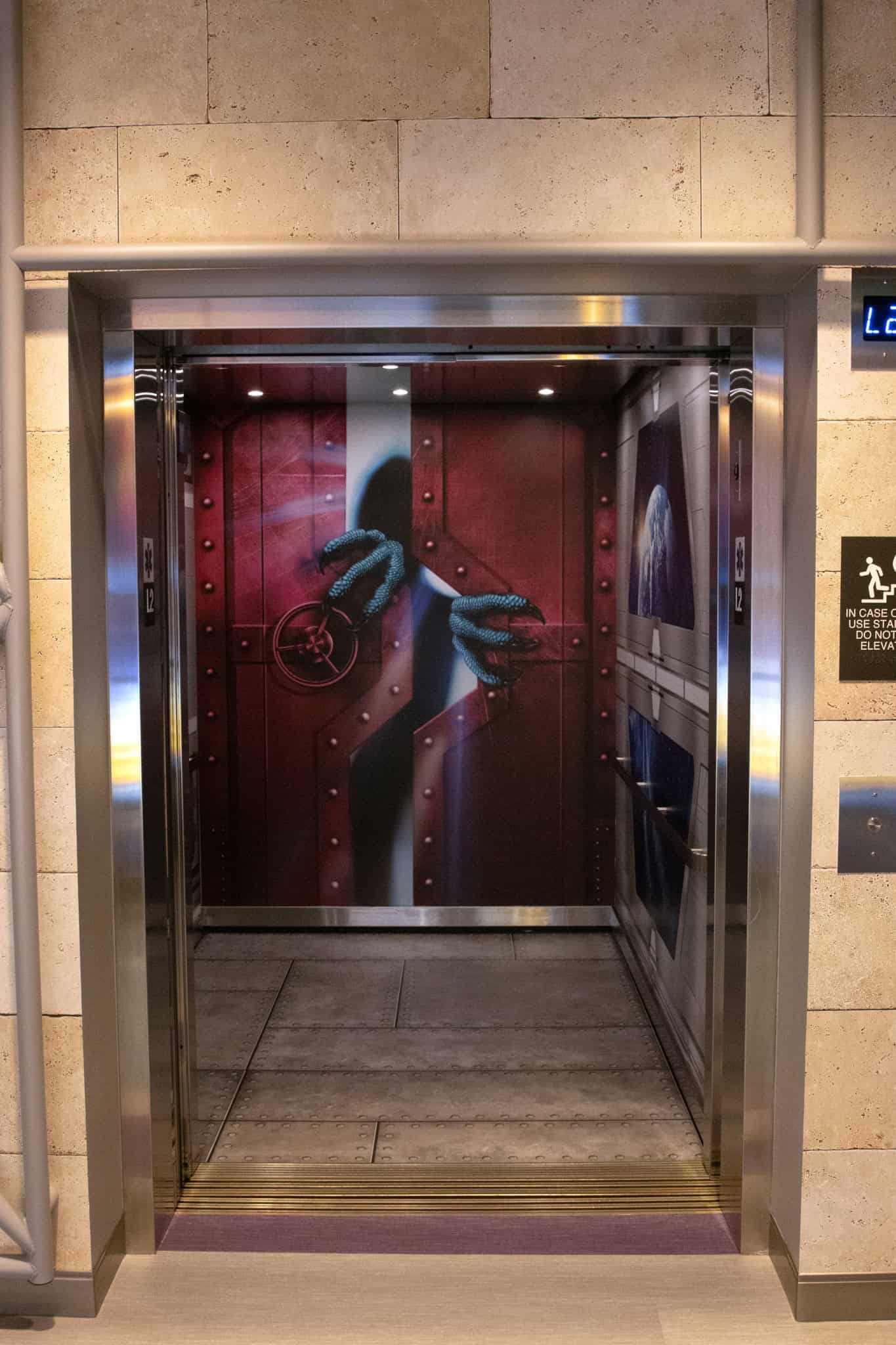 Elevator interior designed like a spaceship, featuring a red containment door with glowing blue monster claws emerging from behind it.