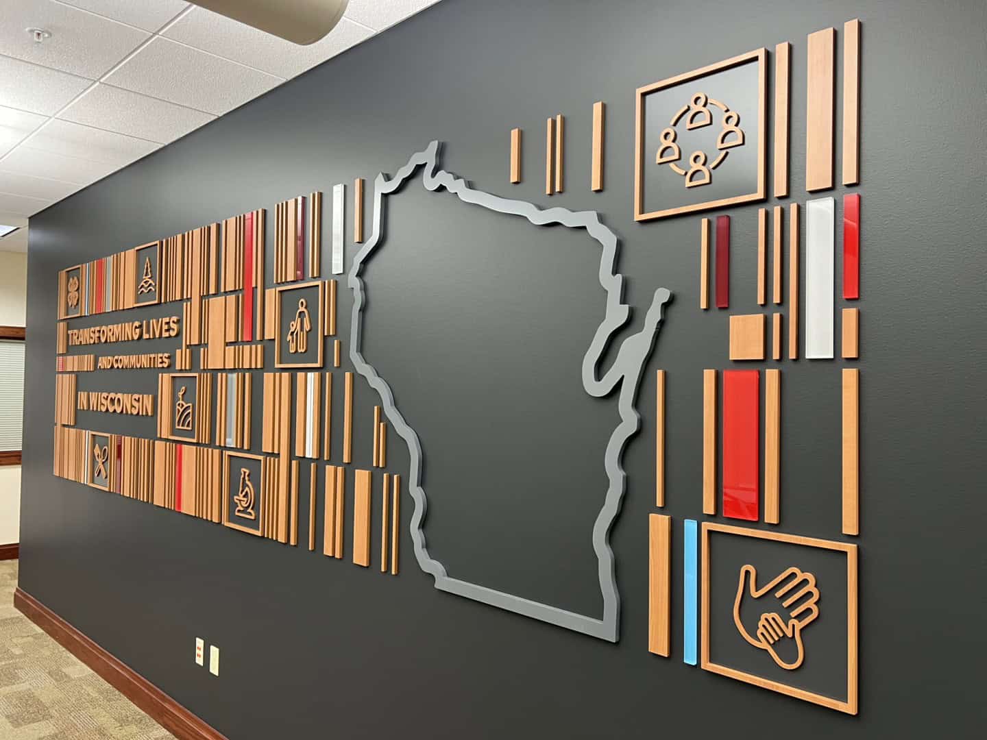 Angled perspective of a bold feature wall with dimensional wood slats and icons, concluding with a large silhouette of Wisconsin and outreach-themed illustrations.