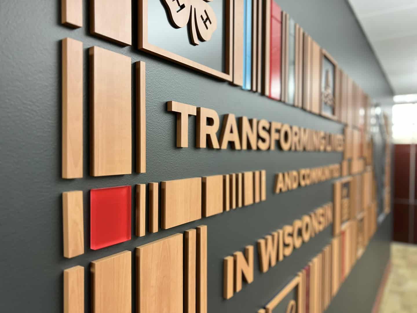 Close-up of a dimensional wall mural with raised wood blocks and text that reads “Transforming Lives and Communities in Wisconsin,” with pops of red and blue acrylic.
