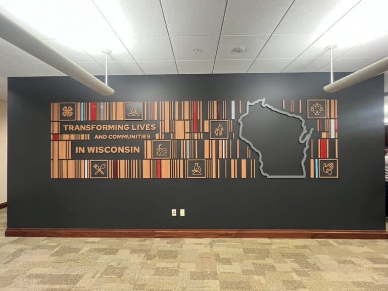 Full view of the dimensional feature wall at UW Extension, displaying wooden graphic elements, icons, and the message “Transforming Lives and Communities in Wisconsin” next to a large outline of the state.