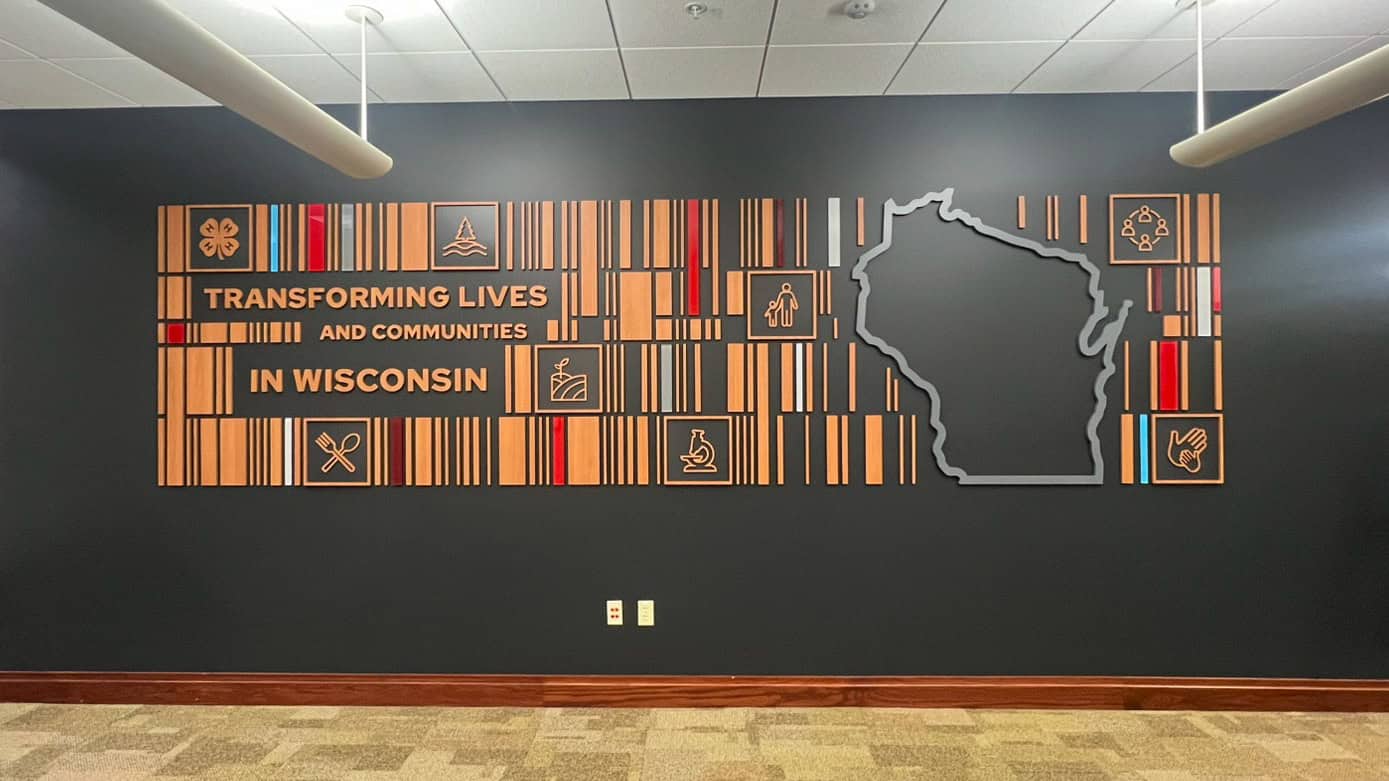 Full view of the dimensional feature wall at UW Extension, displaying wooden graphic elements, icons, and the message “Transforming Lives and Communities in Wisconsin” next to a large outline of the state.