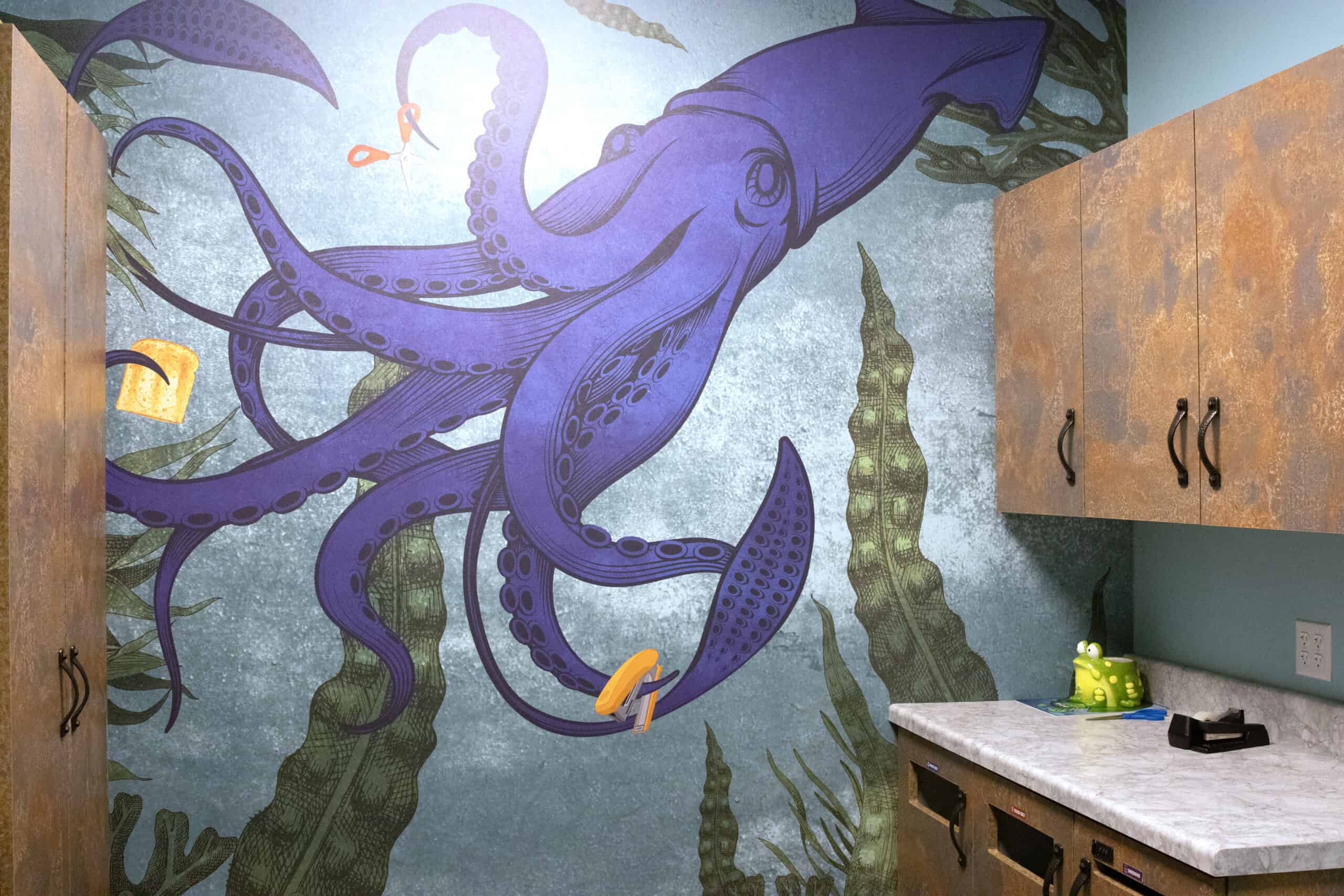Illustrated wall mural featuring a whimsical purple squid holding office supplies with its tentacles, set in a stylized underwater breakroom scene with seaweed and cabinetry.