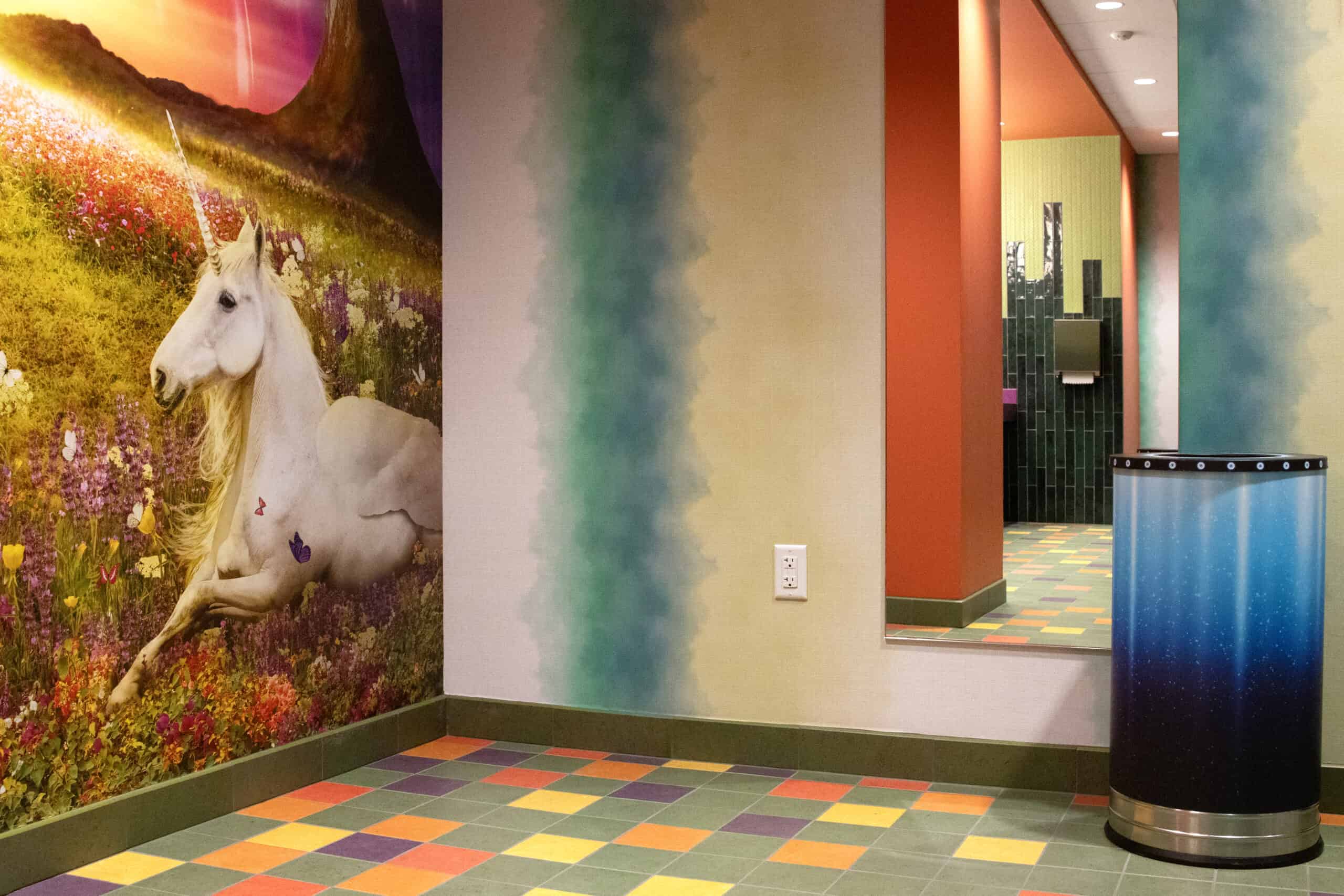 A whimsical wall mural of a unicorn lounging in a flower-filled meadow, next to a multicolored tile floor and gradient-painted walls in a company restroom.