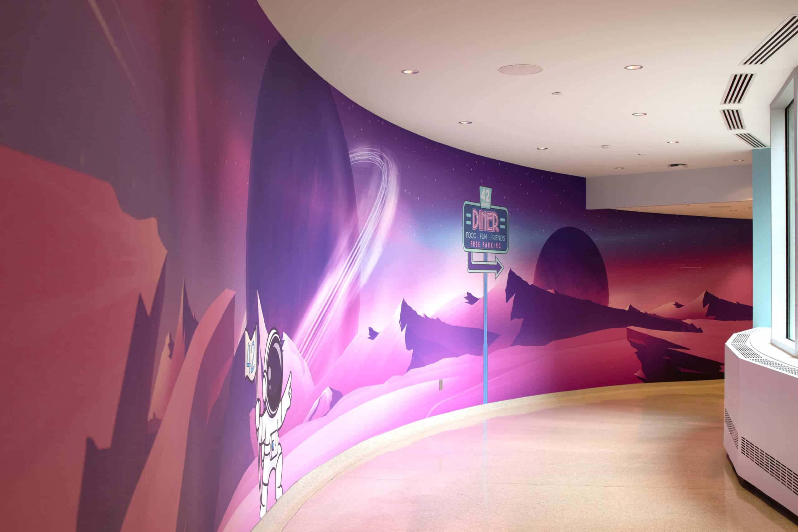 A curved hallway mural depicting a pastel-hued alien landscape with mountains, an astronaut, and a “Diner” sign pointing the way to a themed area.