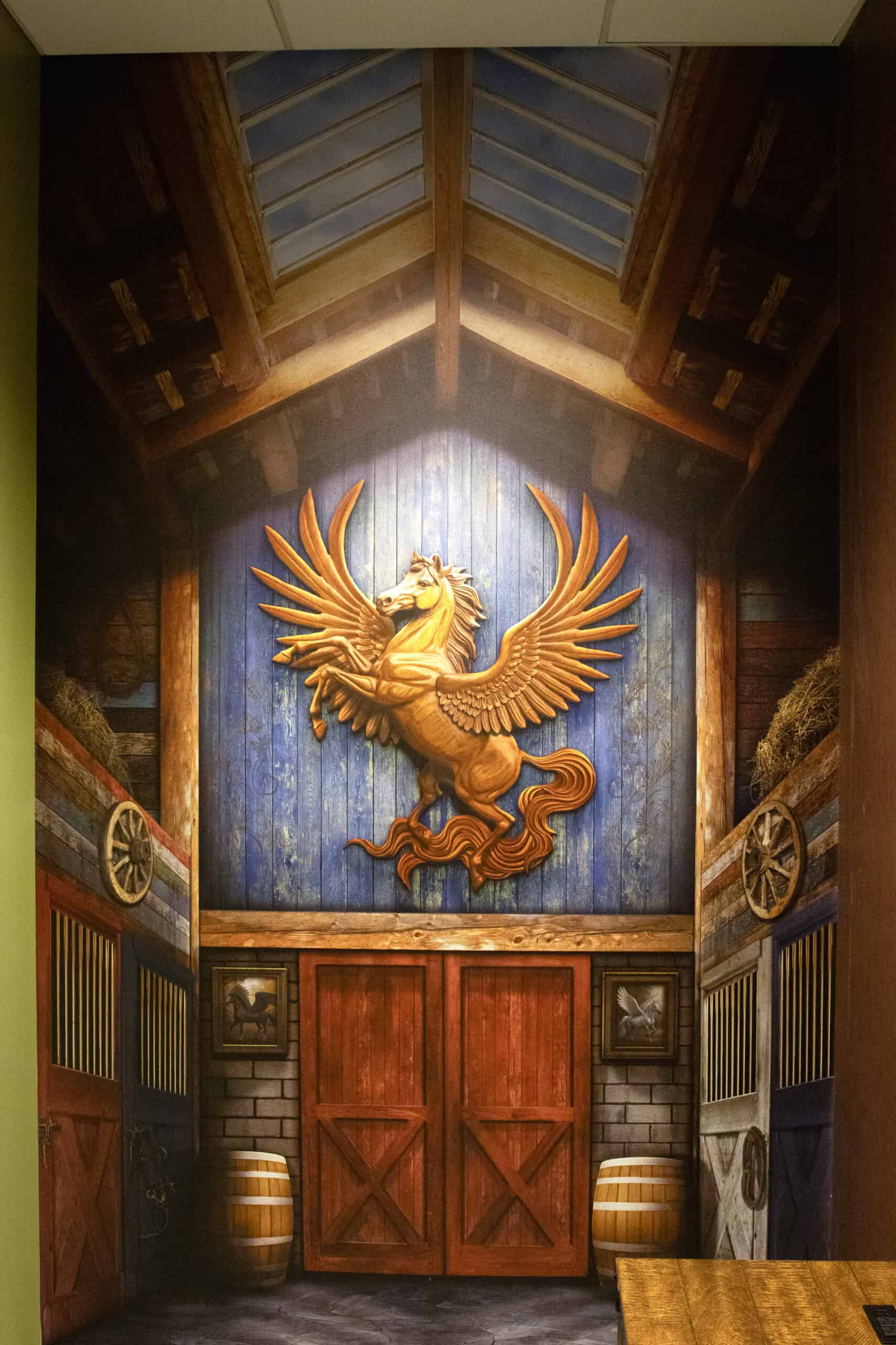 A hyper-realistic mural inside a barn-themed room, with a sculpted Pegasus above wooden stall doors and rustic detailing.