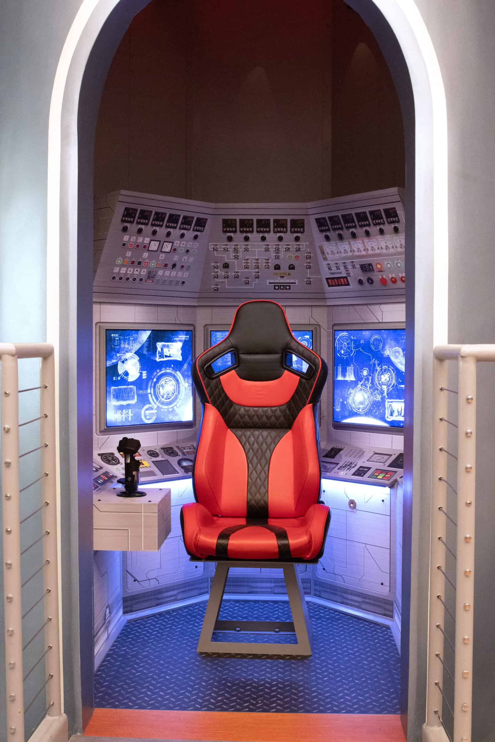 Themed room resembling a spaceship control panel, complete with detailed graphic dashboards and a red-and-black gaming chair in the center.