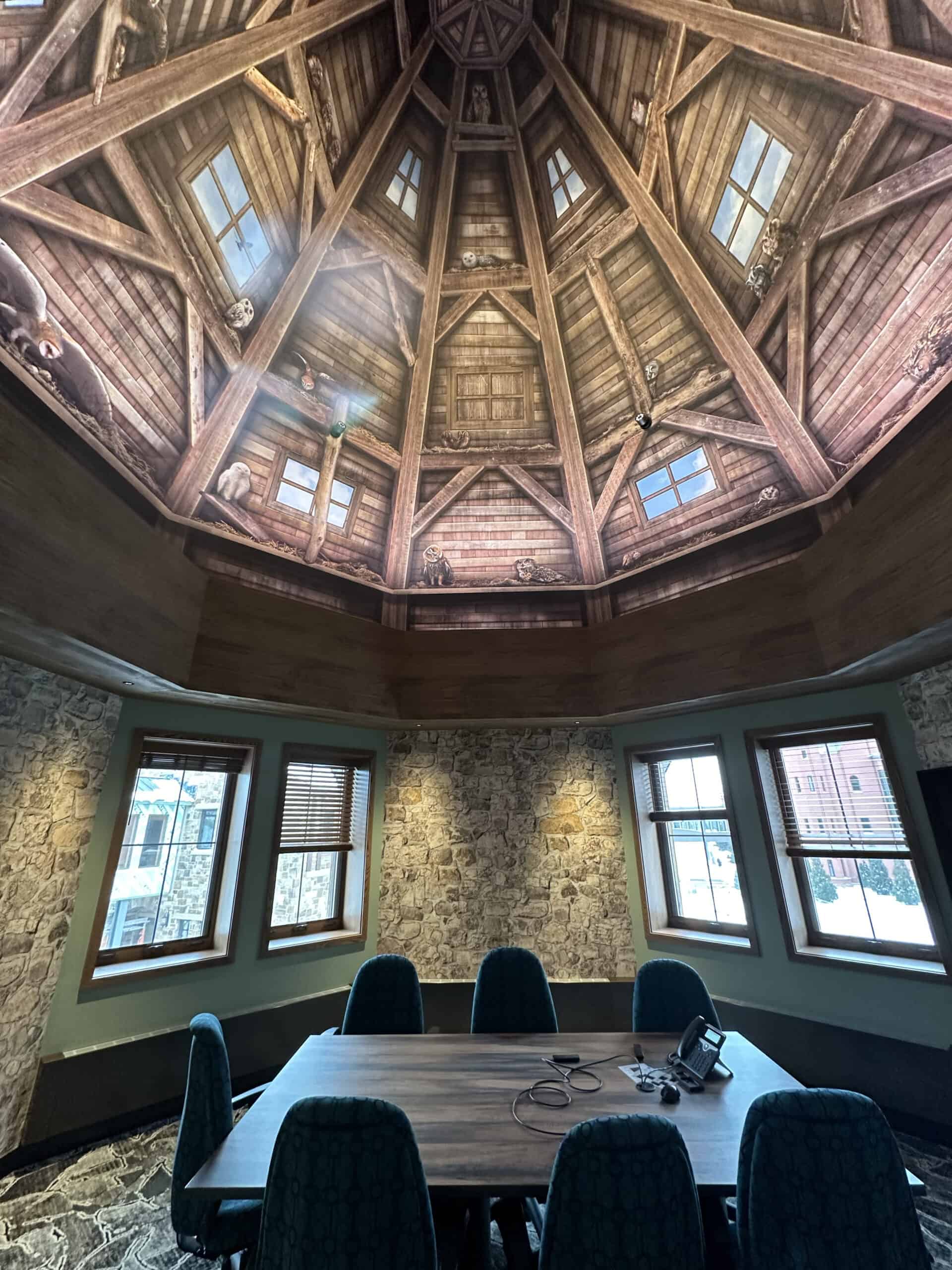 A rustic conference room with sloped stone walls and panoramic windows, featuring a vaulted ceiling mural designed to look like the interior of a wooden turret, complete with illustrated animals tucked into the rafters.