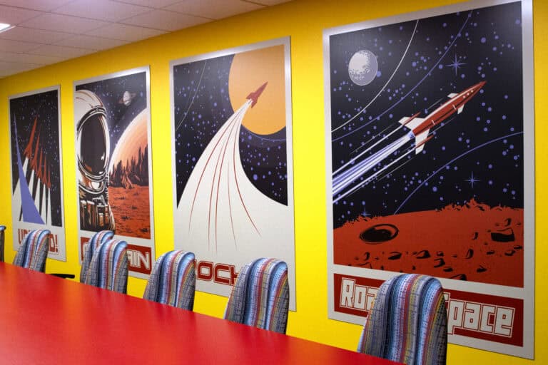 A retro-futuristic themed room featuring bold, graphic wall posters of rockets and astronauts set against a bright yellow wall, with a red conference table and striped multicolor chairs.