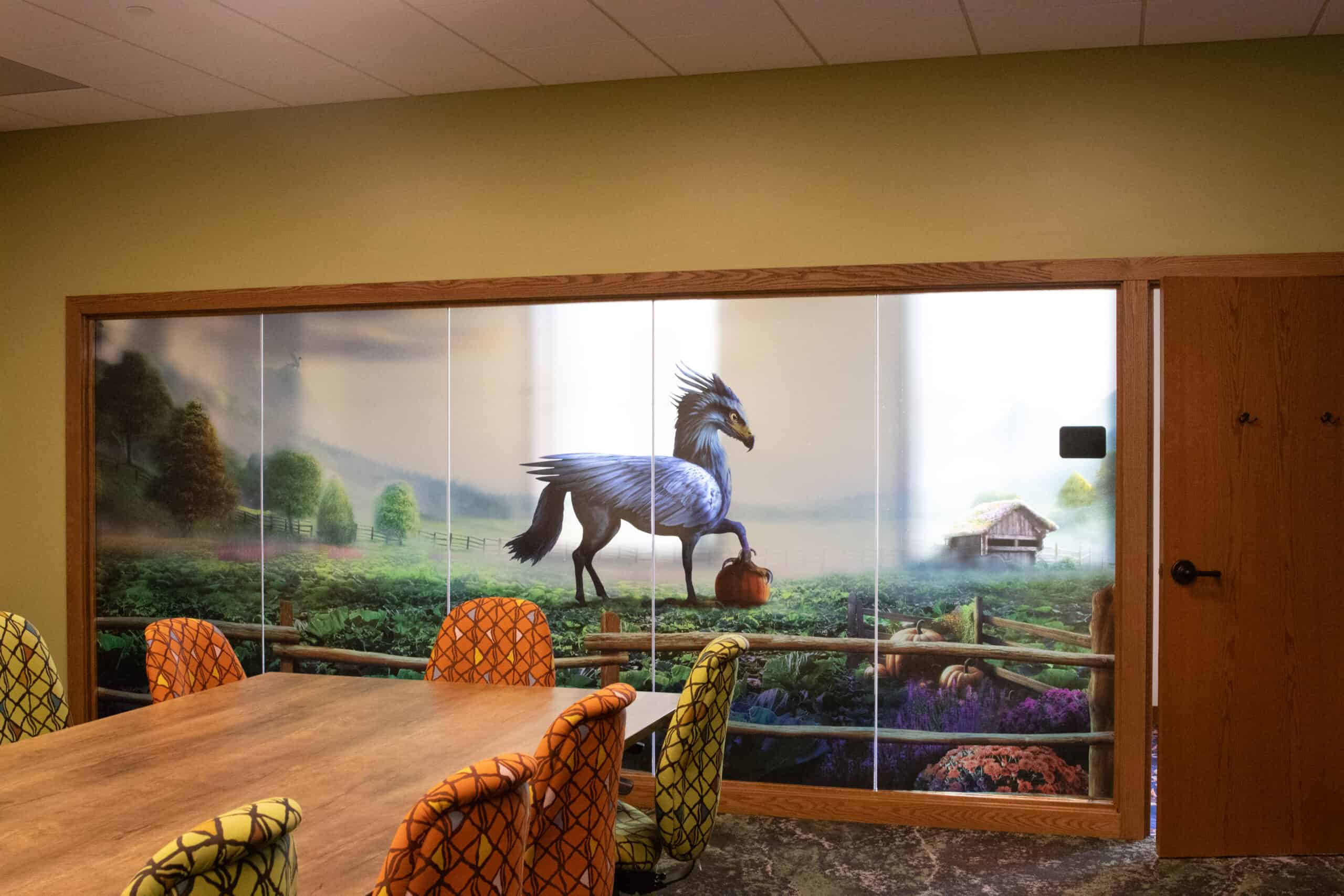 A continuation of the whimsical farmland scene, this room shows a blue mythical bird-horse hybrid carrying a pumpkin in its beak, set against a foggy mountain and barn backdrop with a wooden table and colorful chairs.