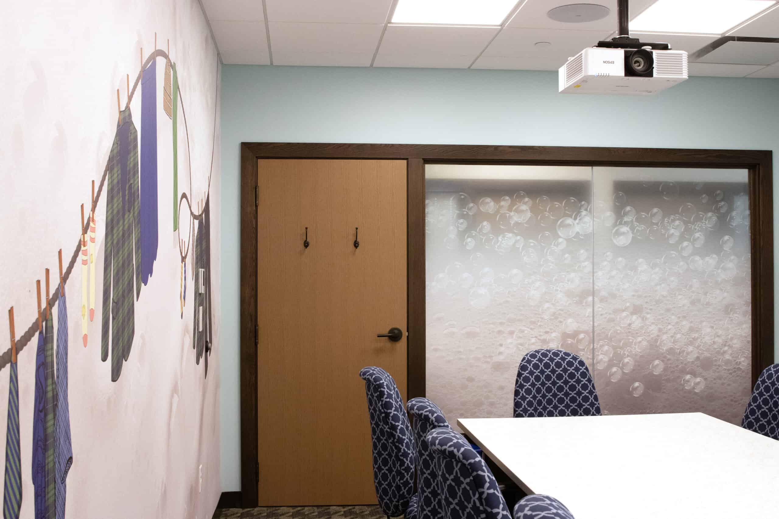 A playful laundry-themed conference room with illustrated clothes hanging on a line along one wall and a bubble-covered frosted window on the opposite side, complemented by navy patterned chairs and pale blue walls.