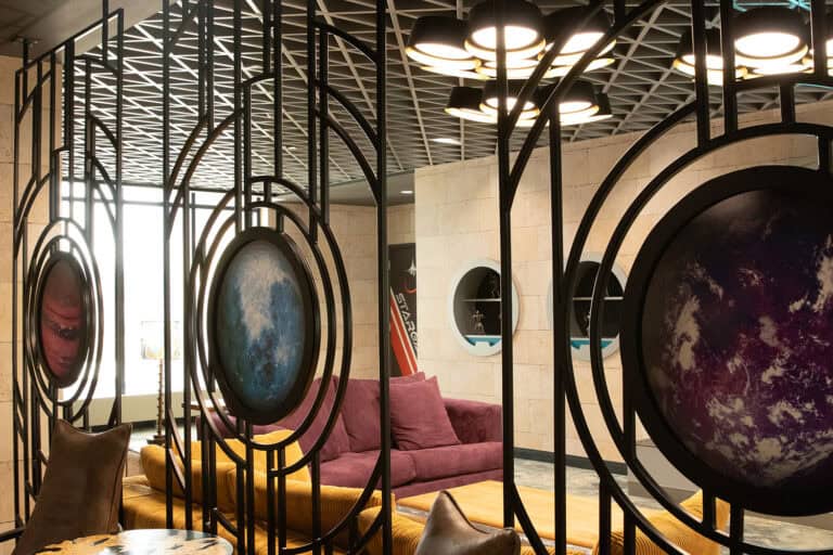 A modern lounge divided by black metal grid partitions framing planet-themed graphics, with cozy seating in purple and yellow tones behind them.