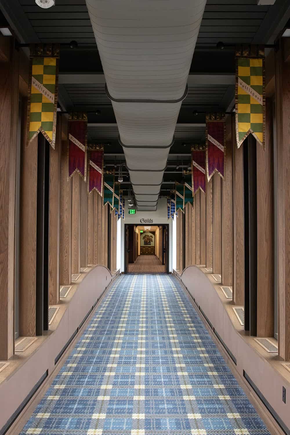 A long, enclosed hallway with plaid carpet, wood paneling, and ceiling banners displaying guild names like Wufflegust and Dragonden, leading to a sign labeled “Guilds.”