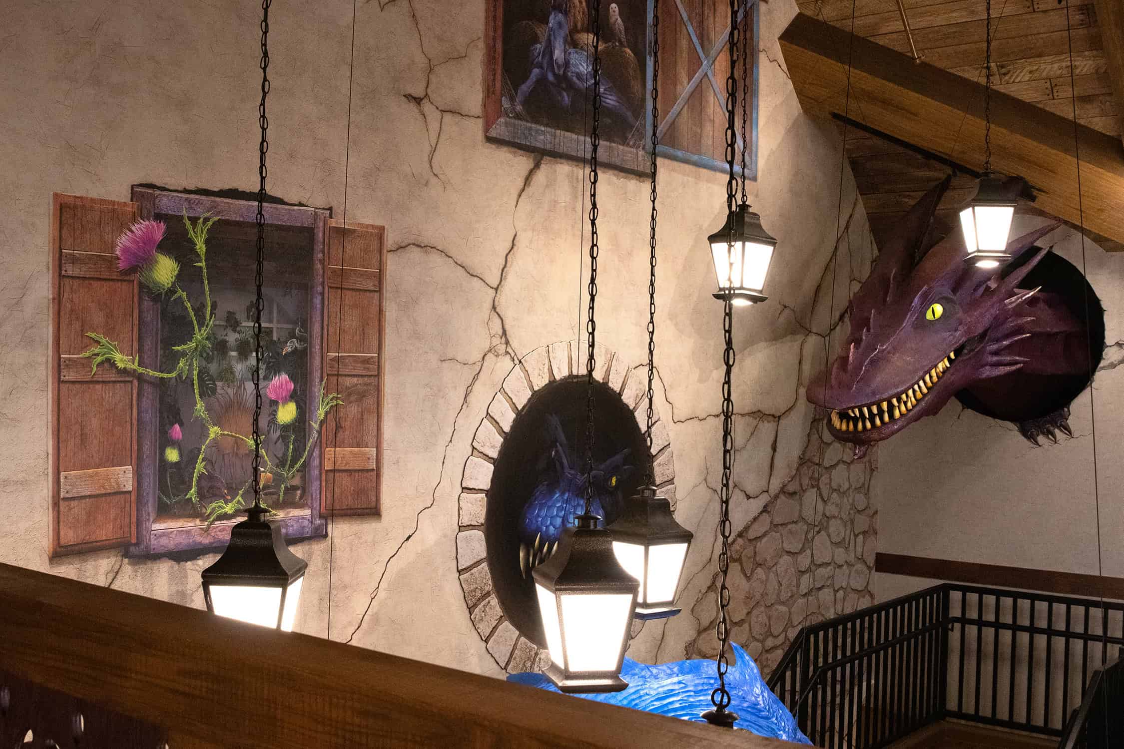 A whimsical stair landing with wall murals depicting castle windows and thistles, with dragon heads emerging from faux stone portals and hanging lanterns adding ambiance.