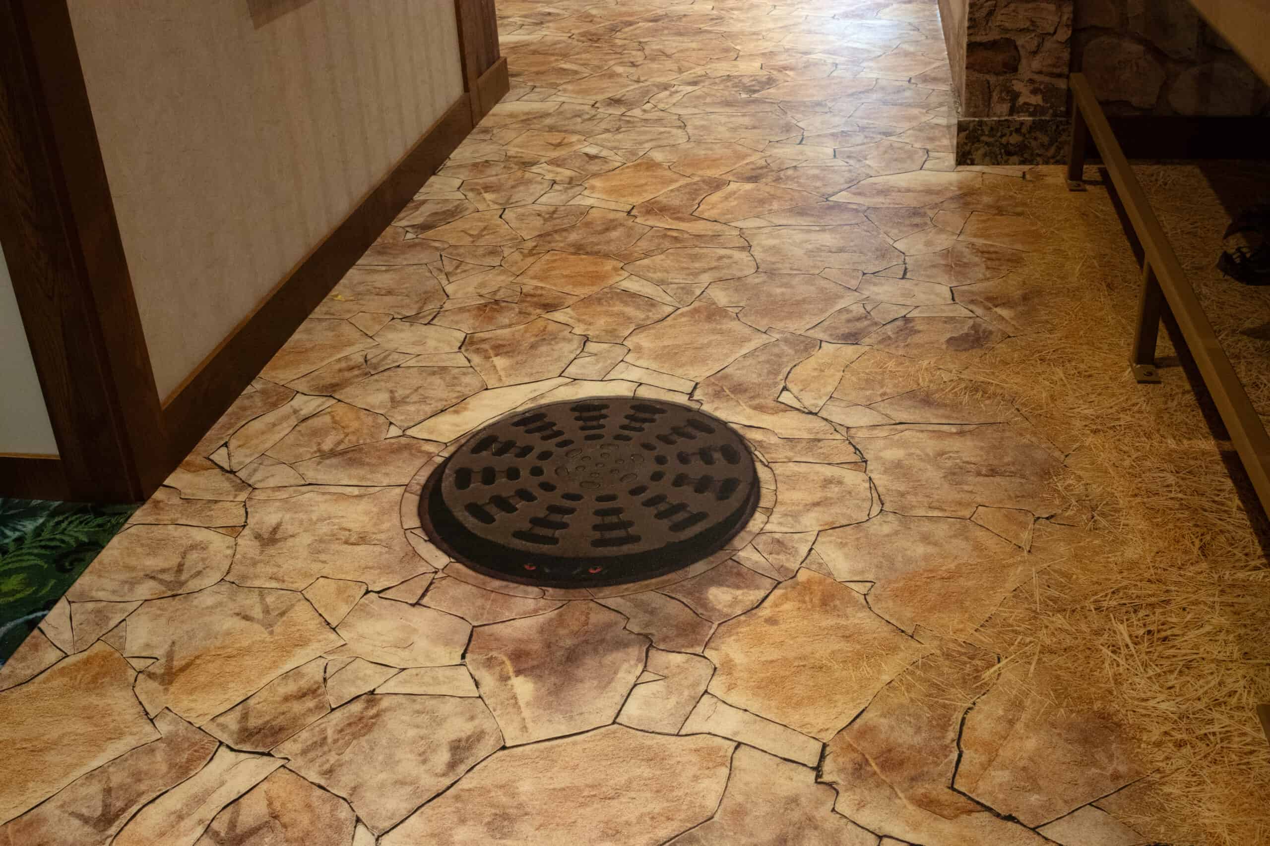 Realistic floor graphic mimicking a stone path with a manhole cover in the center, surrounded by printed hay and animal tracks.