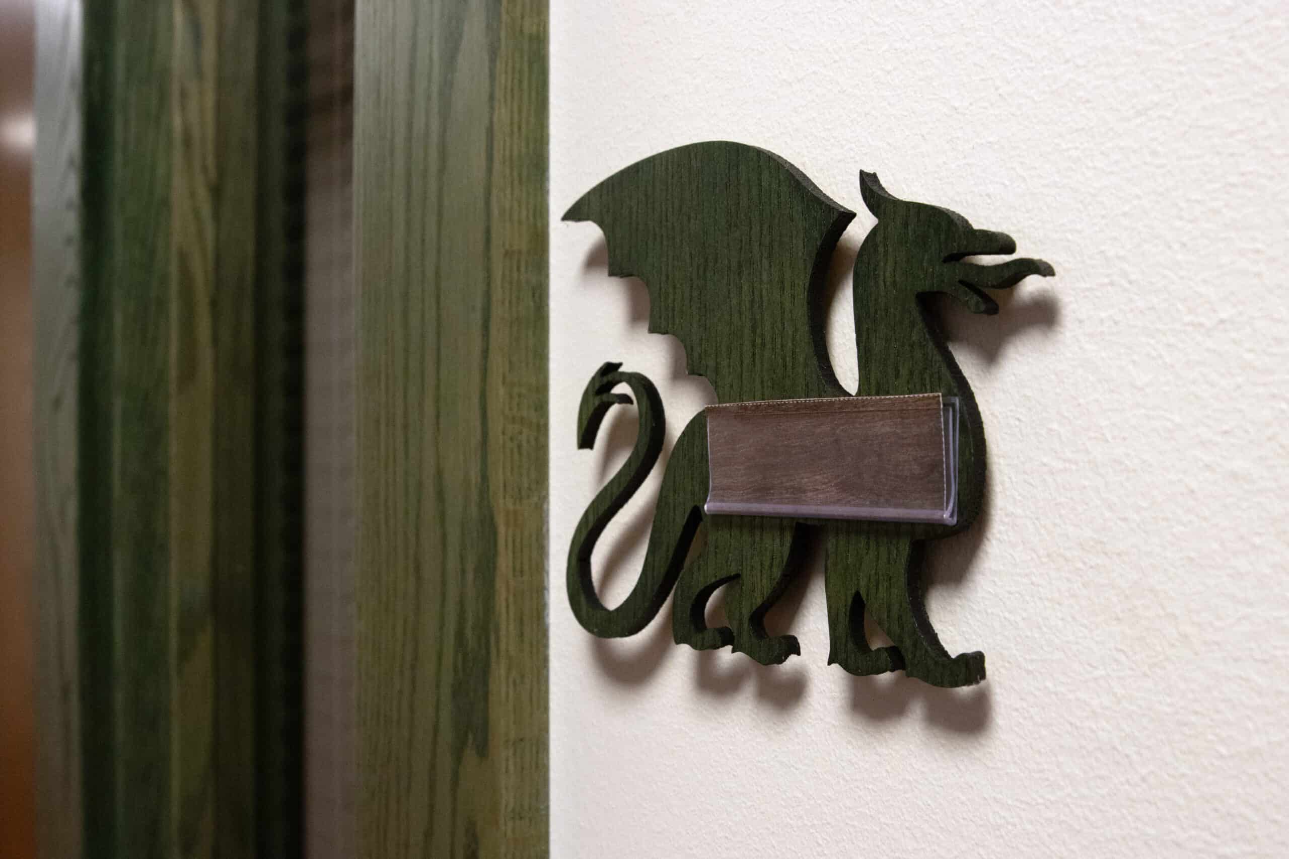 Dark green wood cutout of a dragon used as a room sign, mounted to the wall with a blank nameplate.