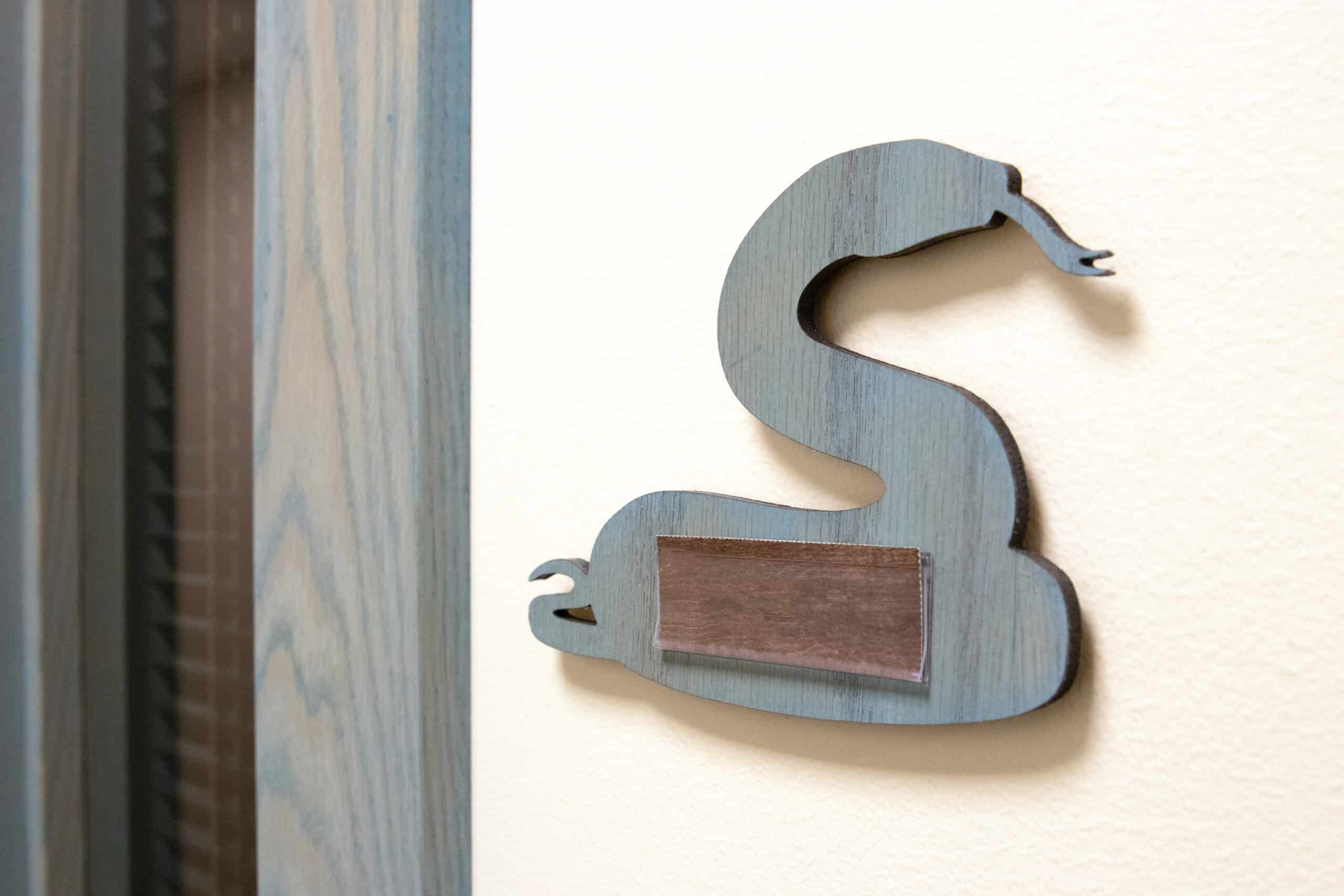 Blue wood cutout of a snake used as a room sign, mounted on the wall with a blank nameplate insert.