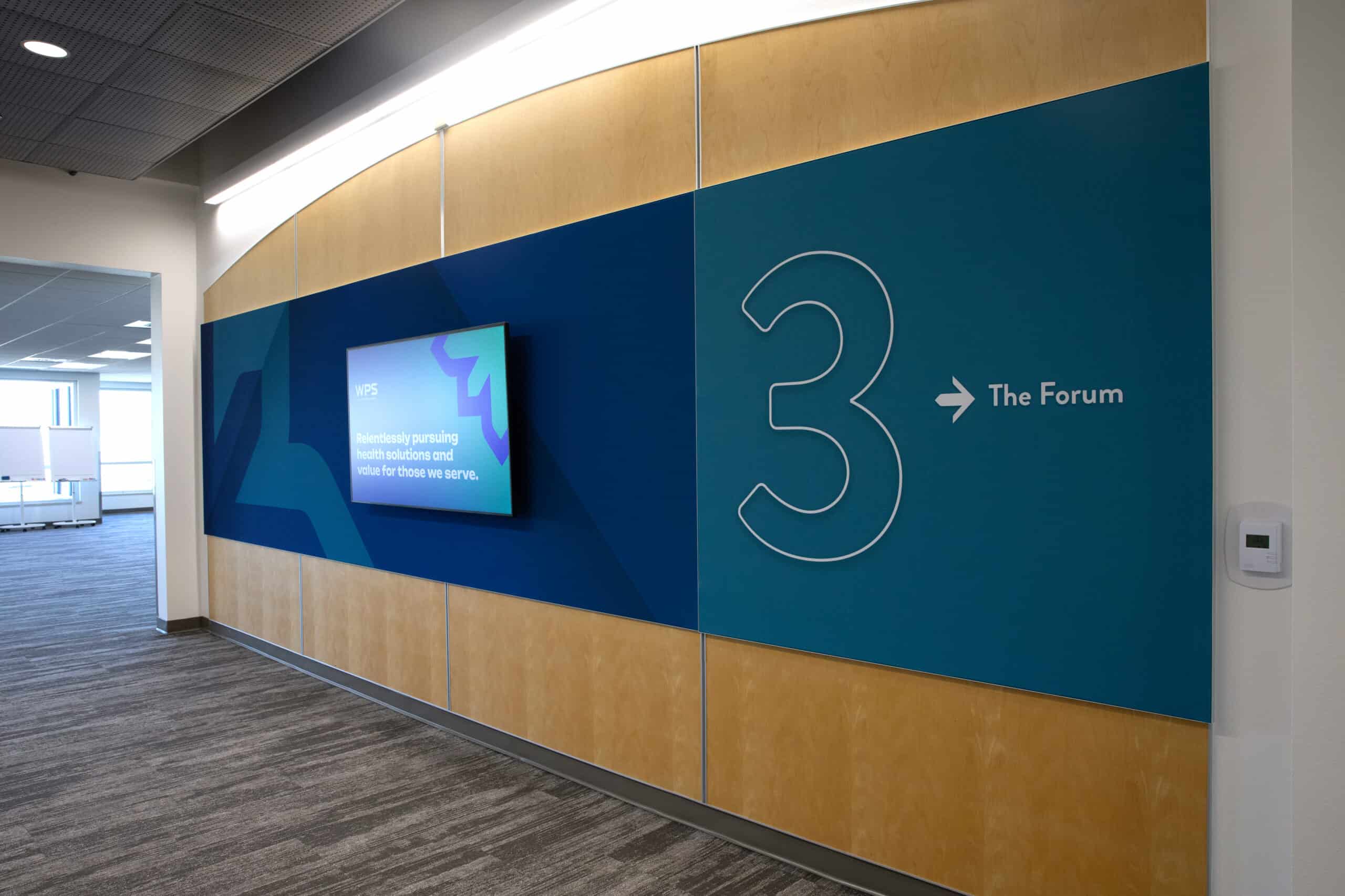 Branded wayfinding wall at WPS headquarters with monitor display and large numeral 3 directing to The Forum meeting space.