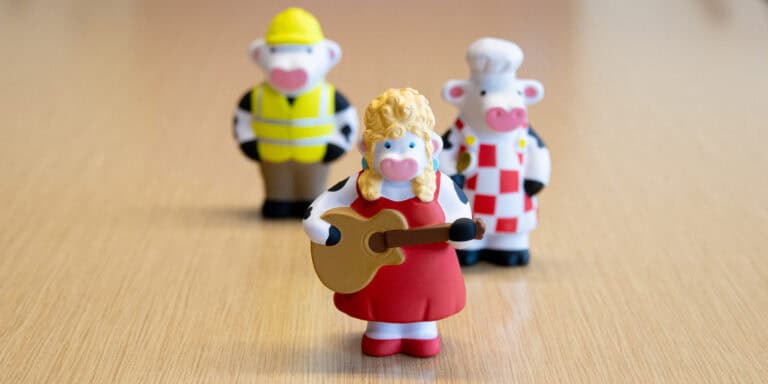 Custom cow figurine dressed as a country music singer with a red dress, blonde curls, and a guitar stands in focus on a woodgrain surface. Behind her are two other cow figurines in soft focus, one dressed as a construction worker and the other as a chef.