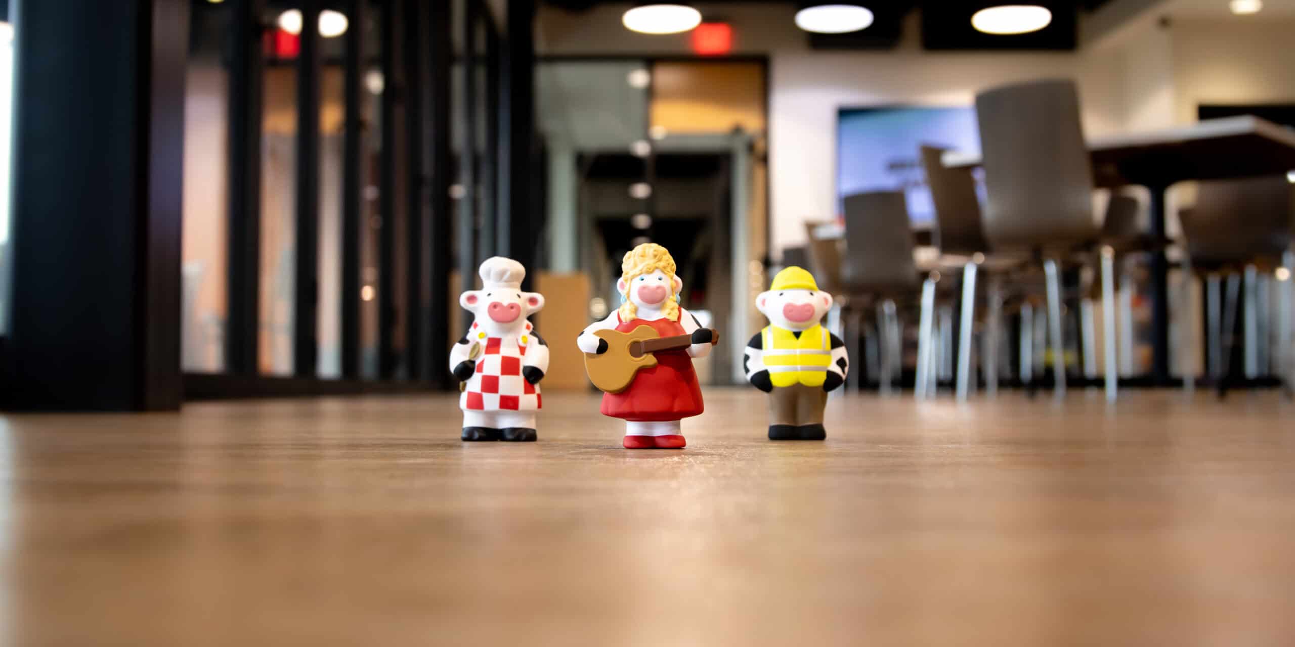 Custom cow figurine dressed as a country music singer with a red dress, blonde curls, and a guitar stands in focus on a woodgrain surface. Behind her are two other cow figurines in soft focus, one dressed as a construction worker and the other as a chef.